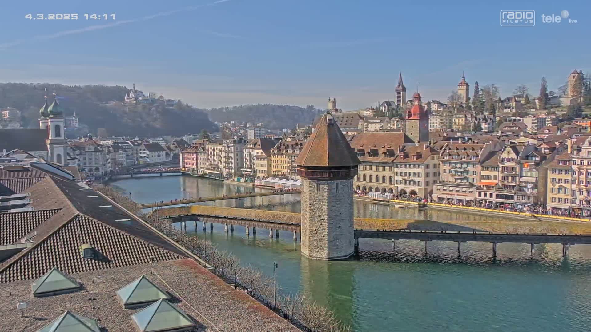 A stone tower stands in the middle of a river, connected to both sides by a long wooden bridge, with a town of colorful buildings on a hill in the background under a clear, sunny sky.