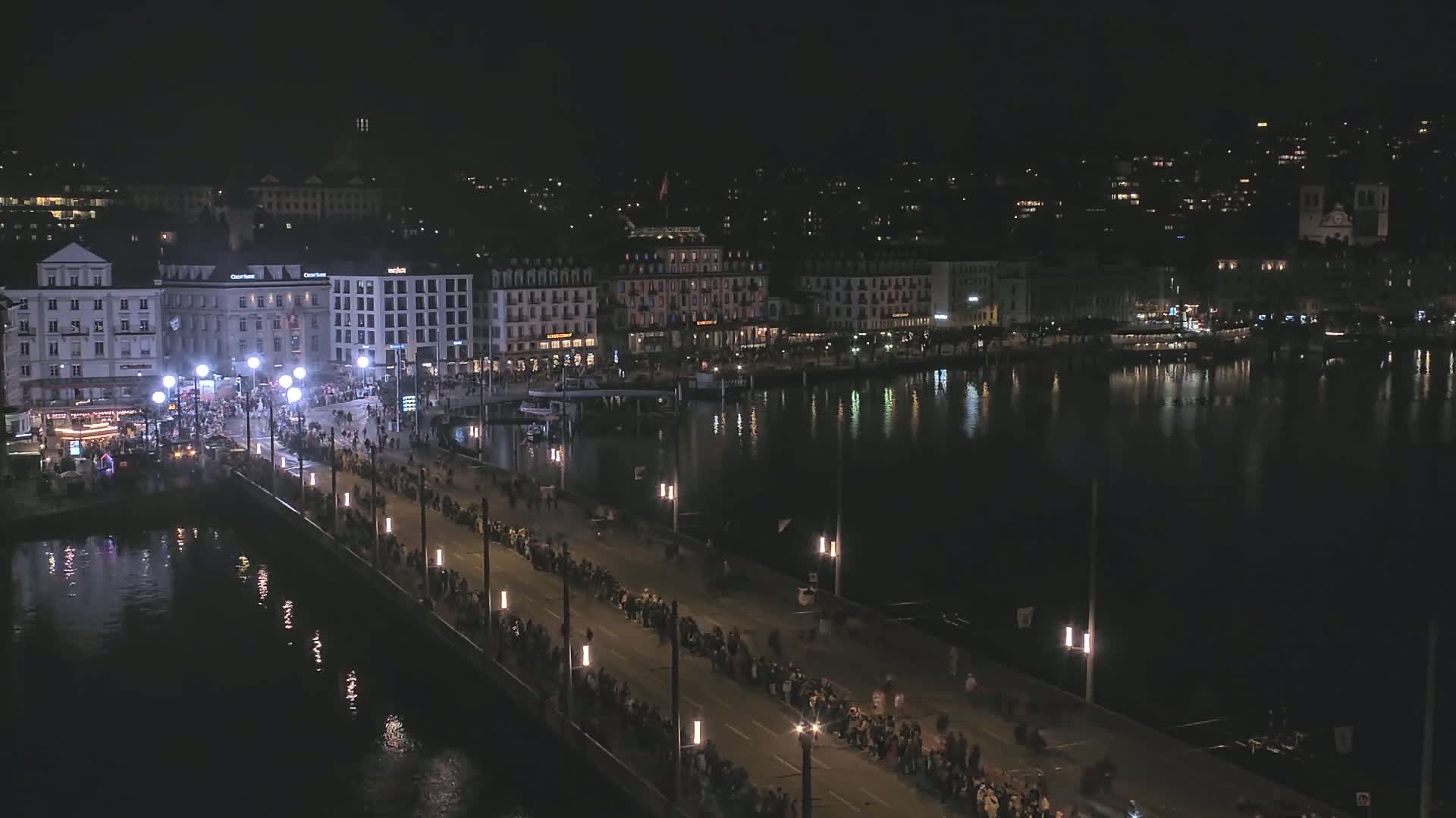 A nighttime, aerial view shows a crowd of people lining a bridge that spans a dark, calm body of water, with city buildings illuminated along the waterfront.