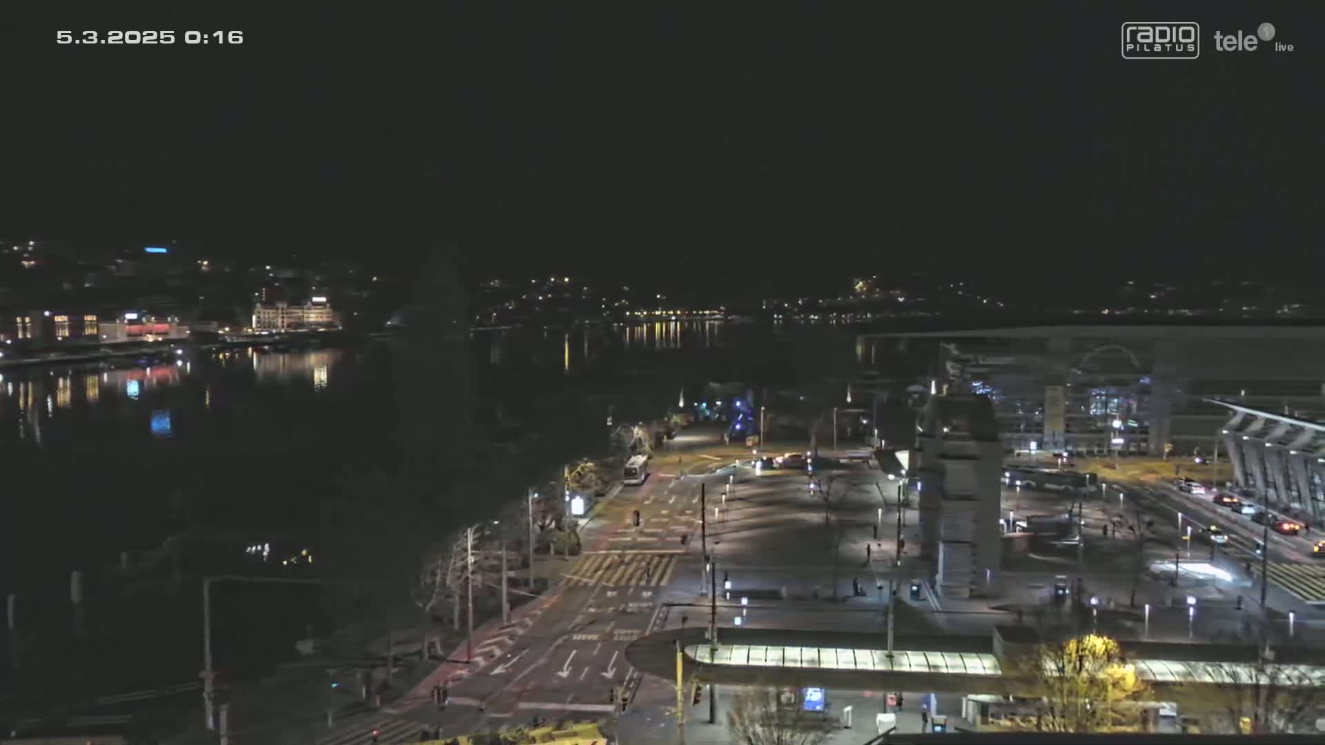 A nighttime, high-angle view shows a city waterfront with calm water reflecting lights from buildings across the water, along with a roadway and city square with sparse traffic.