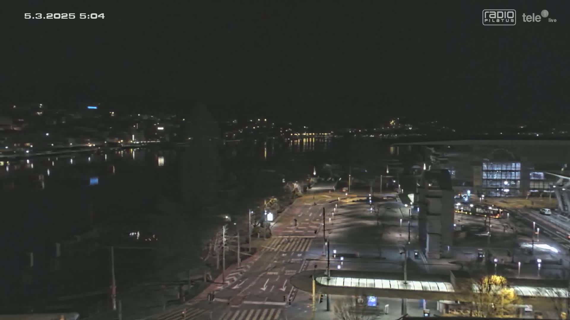 A nighttime, high-angle view shows a city street and waterfront with calm water reflecting city lights.