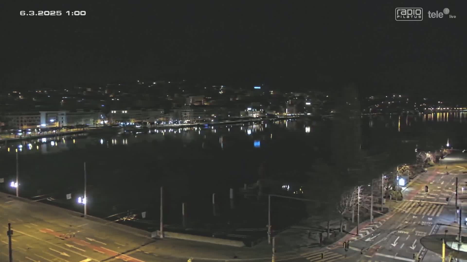 A nighttime view of a city waterfront with calm water reflecting city lights under a clear, dark sky.