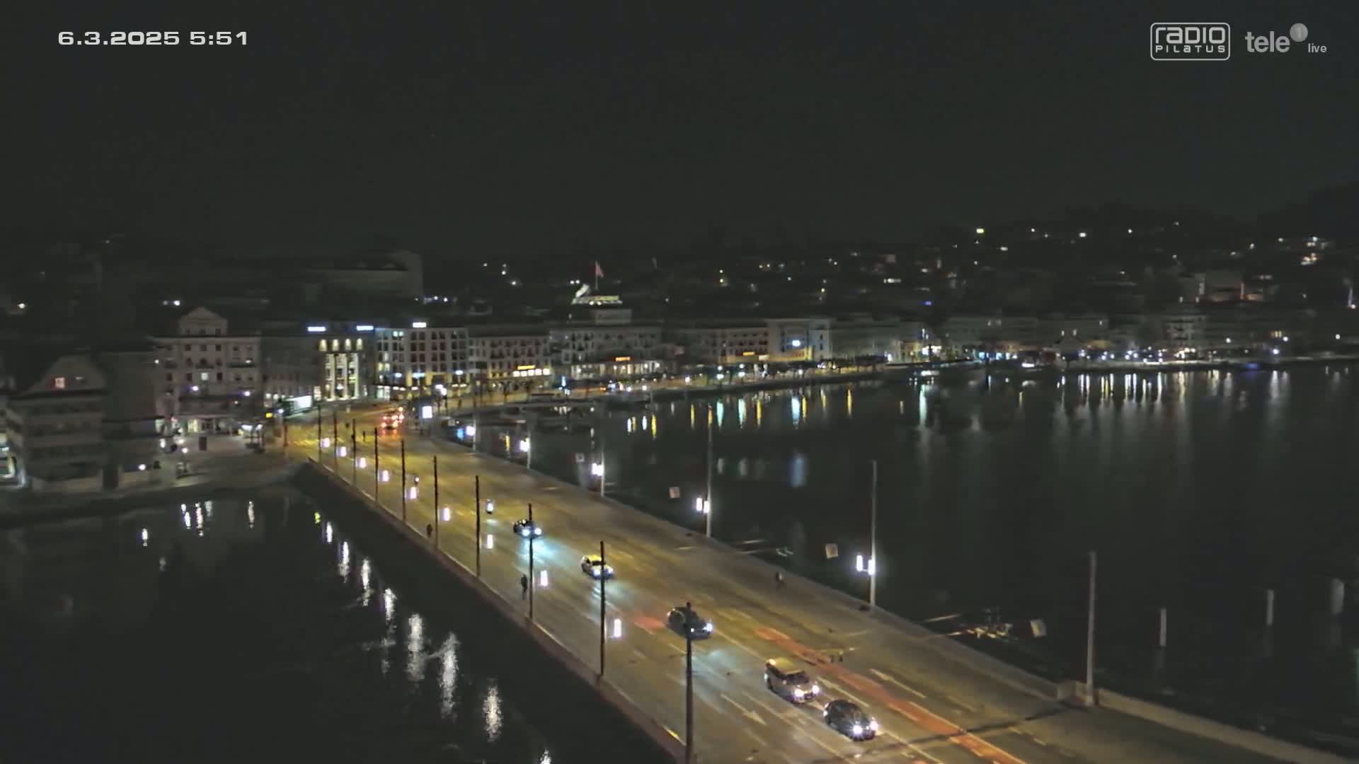 A nighttime, high-angle view shows a bridge with cars driving across it over calm water, next to a city with illuminated buildings.