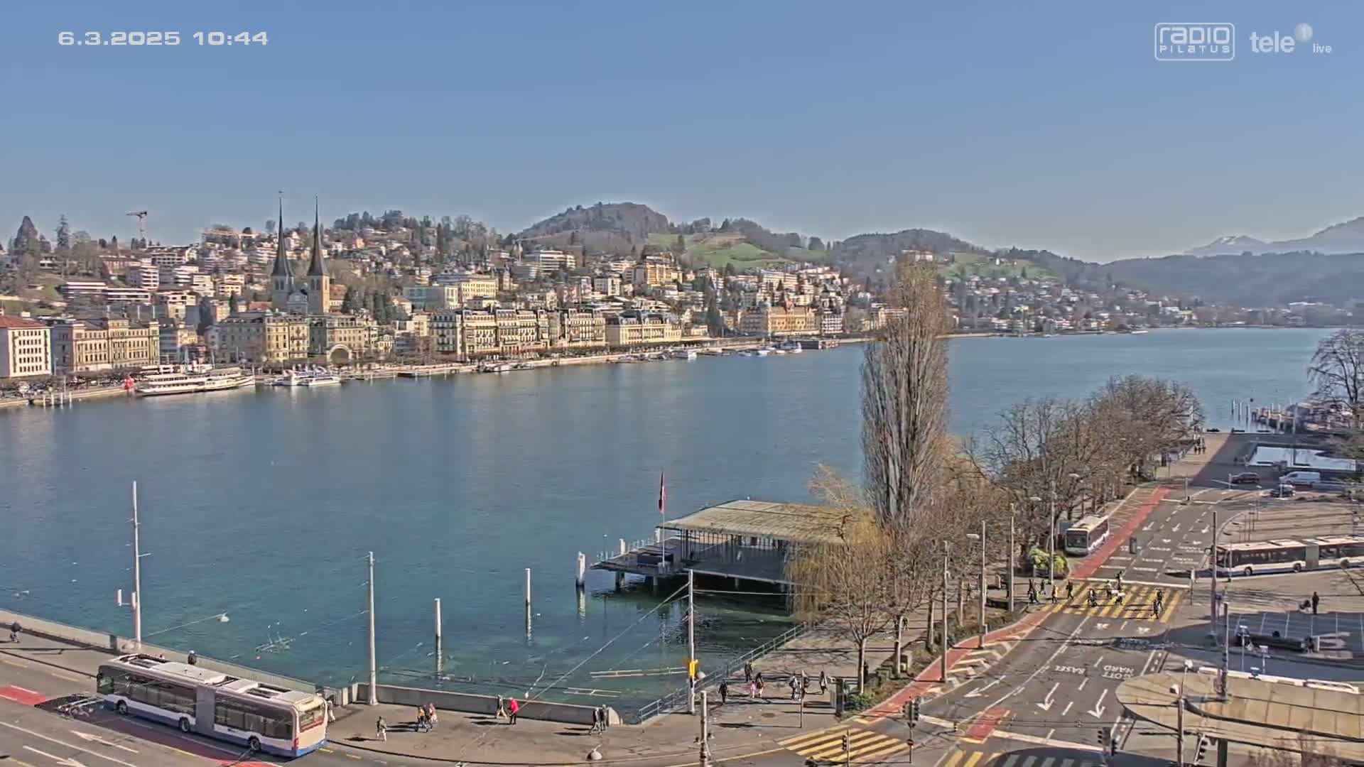 A clear, sunny day shows a waterfront city with buildings lining a calm lake, boats docked, and a roadway with pedestrian crossings.