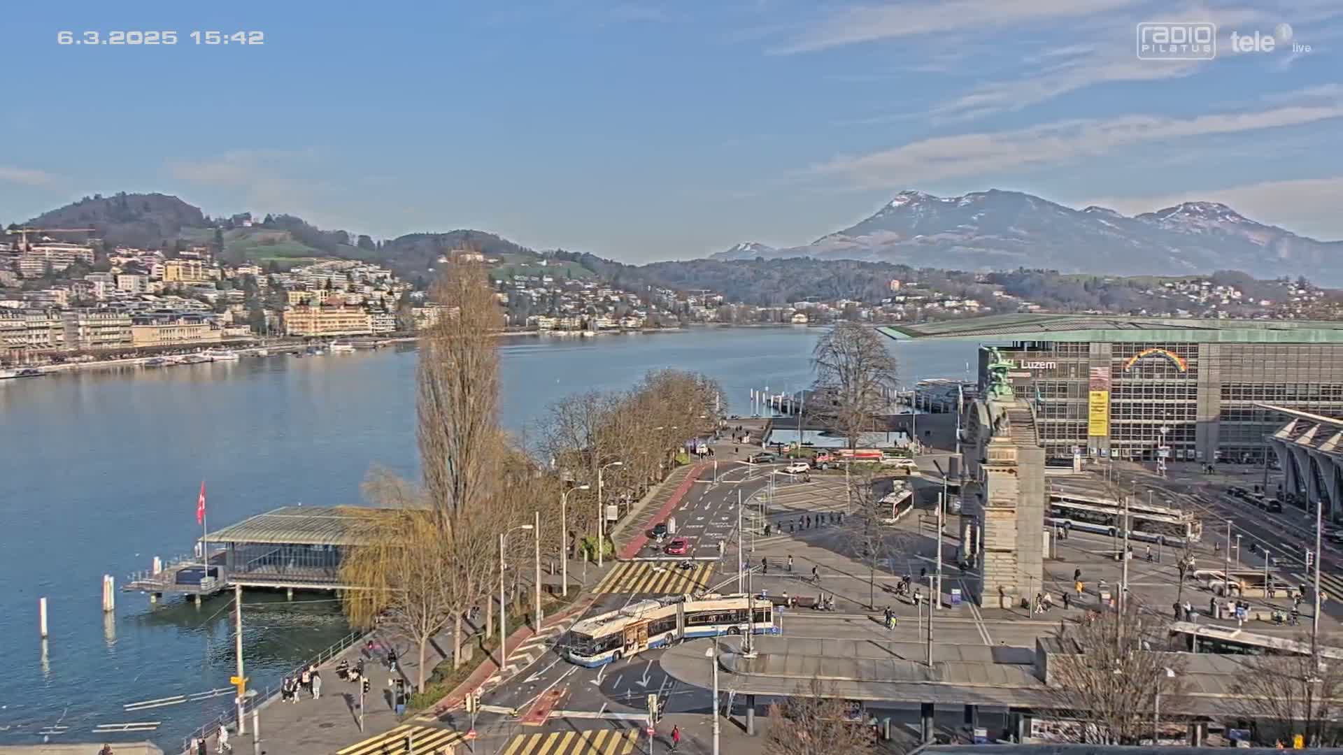 A sunny view of a city by a lake, with snow-capped mountains in the background, showing a waterfront area with buildings, roads, and a trolleybus.