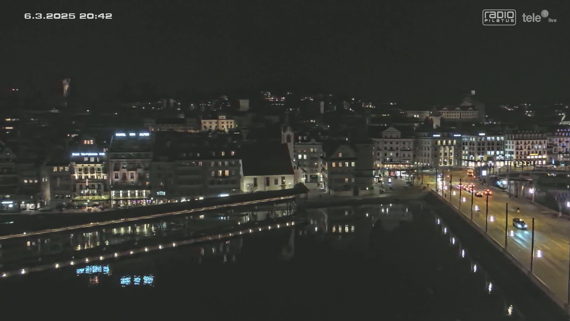 A nighttime aerial view of a city with calm water reflecting the lights of buildings and a bridge, under a clear, dark sky.