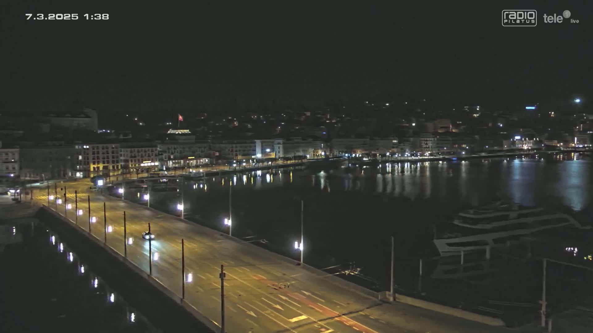 A nighttime view of a city's waterfront, showing a bridge with sparse traffic over calm water alongside docked boats and buildings lit up along the shore.