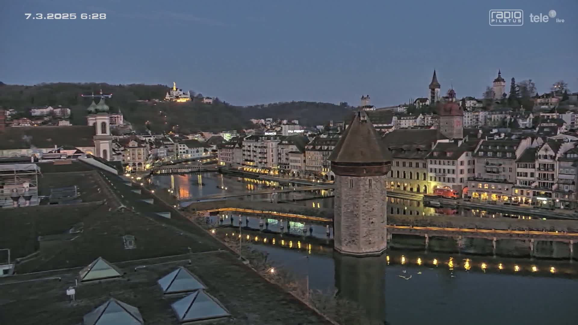 A twilight view of a city with a river, a stone tower in the water, and buildings illuminated along the waterfront.