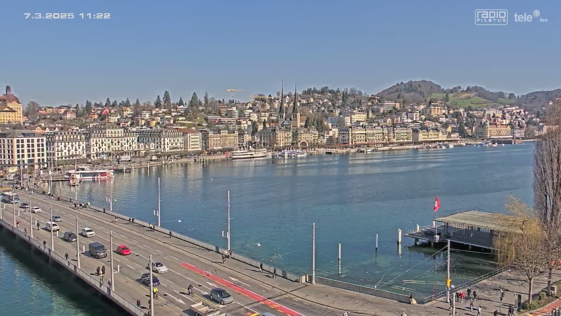 A bridge over calm blue water leads to a city of buildings lining a waterfront on a clear, sunny day.