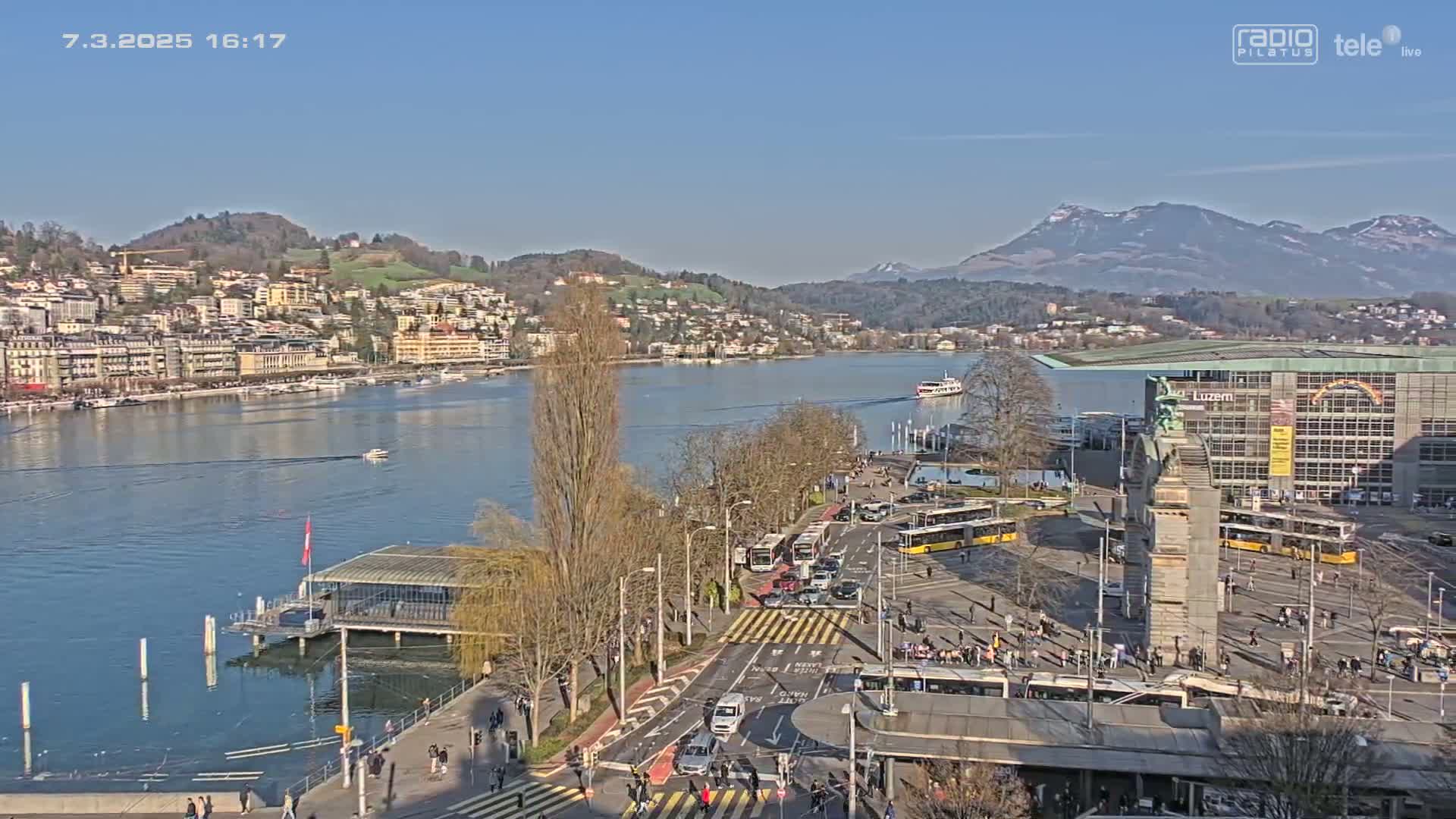 A sunny day overlooking a lake city with mountains in the background, showing a waterfront area with boats, buildings, and buses in a large plaza.