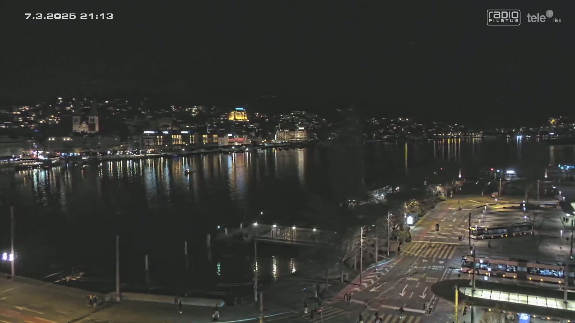 A nighttime, waterfront view of a city with calm water reflecting the lights of buildings and vehicles on a roadway.