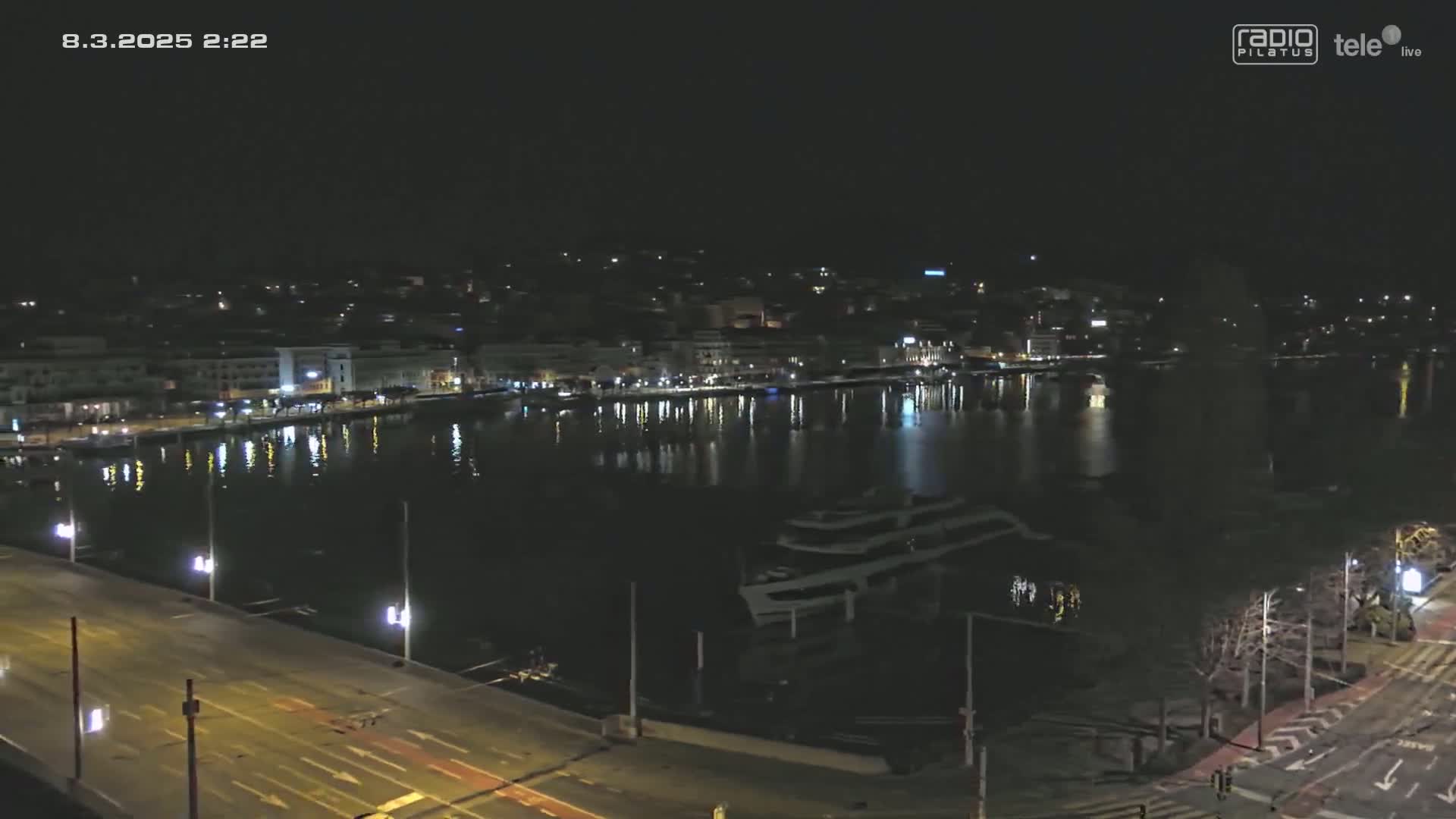 A nighttime view of a city waterfront, showing a calm body of water reflecting the city lights, with a road and streetlights visible in the foreground.