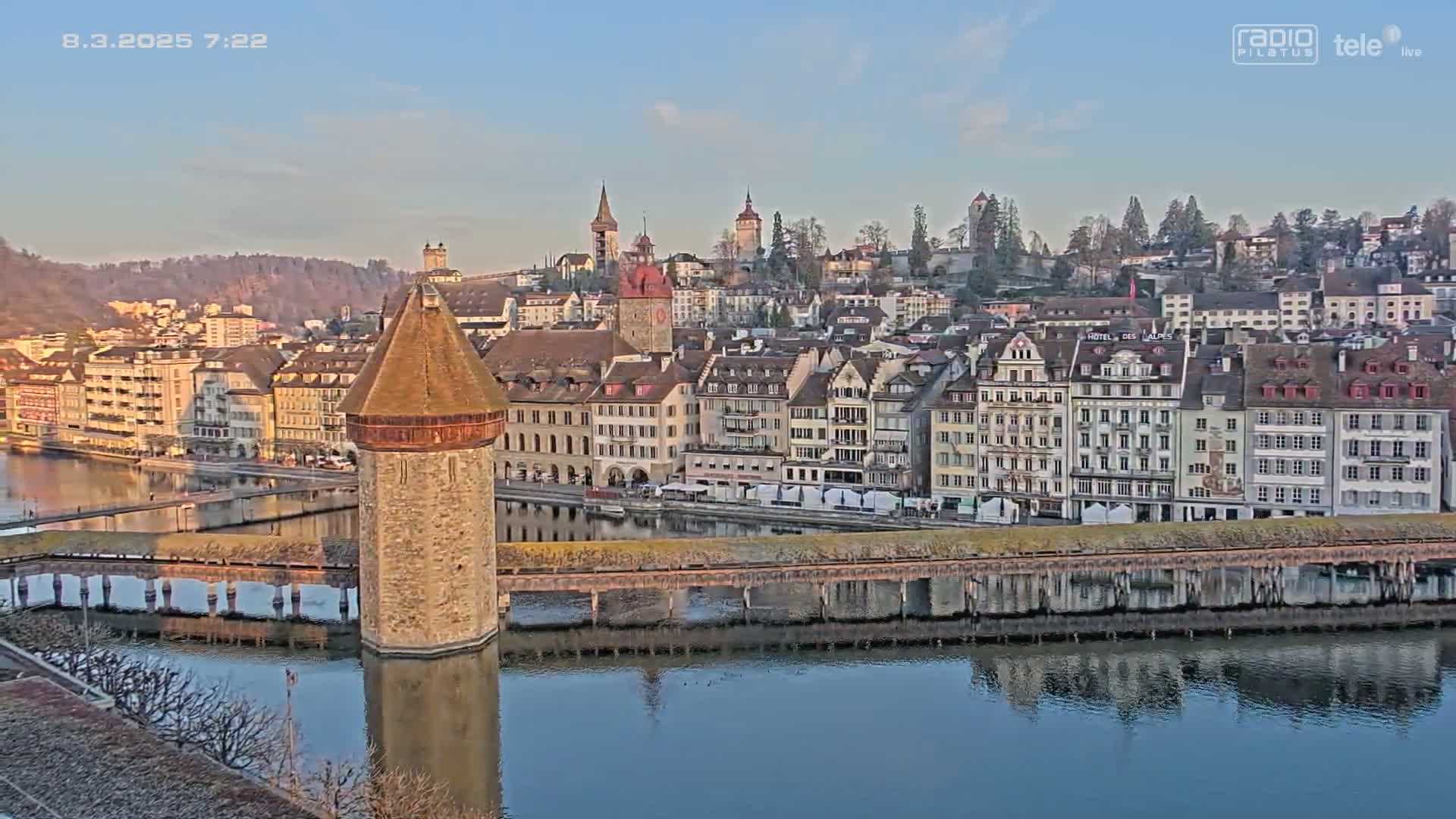 A stone tower stands on a bridge spanning a calm river, reflecting a cityscape of European-style buildings under a clear, early morning sky.