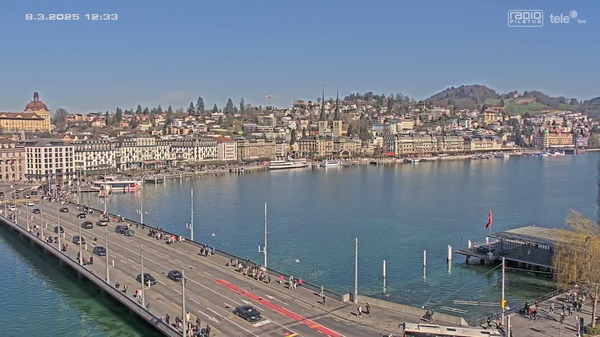 A city waterfront on a sunny day, showing a bridge with cars and pedestrians, boats on the water, and buildings on a hillside.