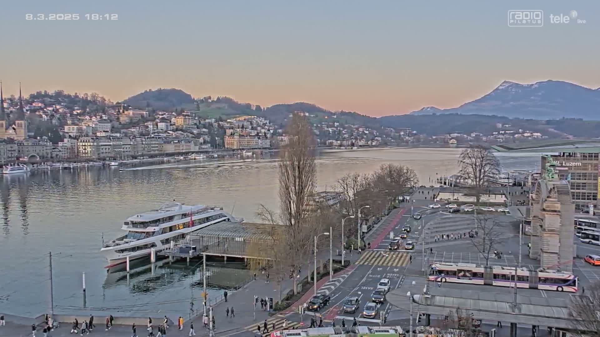 A large passenger ship is docked at a waterfront city during a calm, twilight evening, with distant mountains in the background.