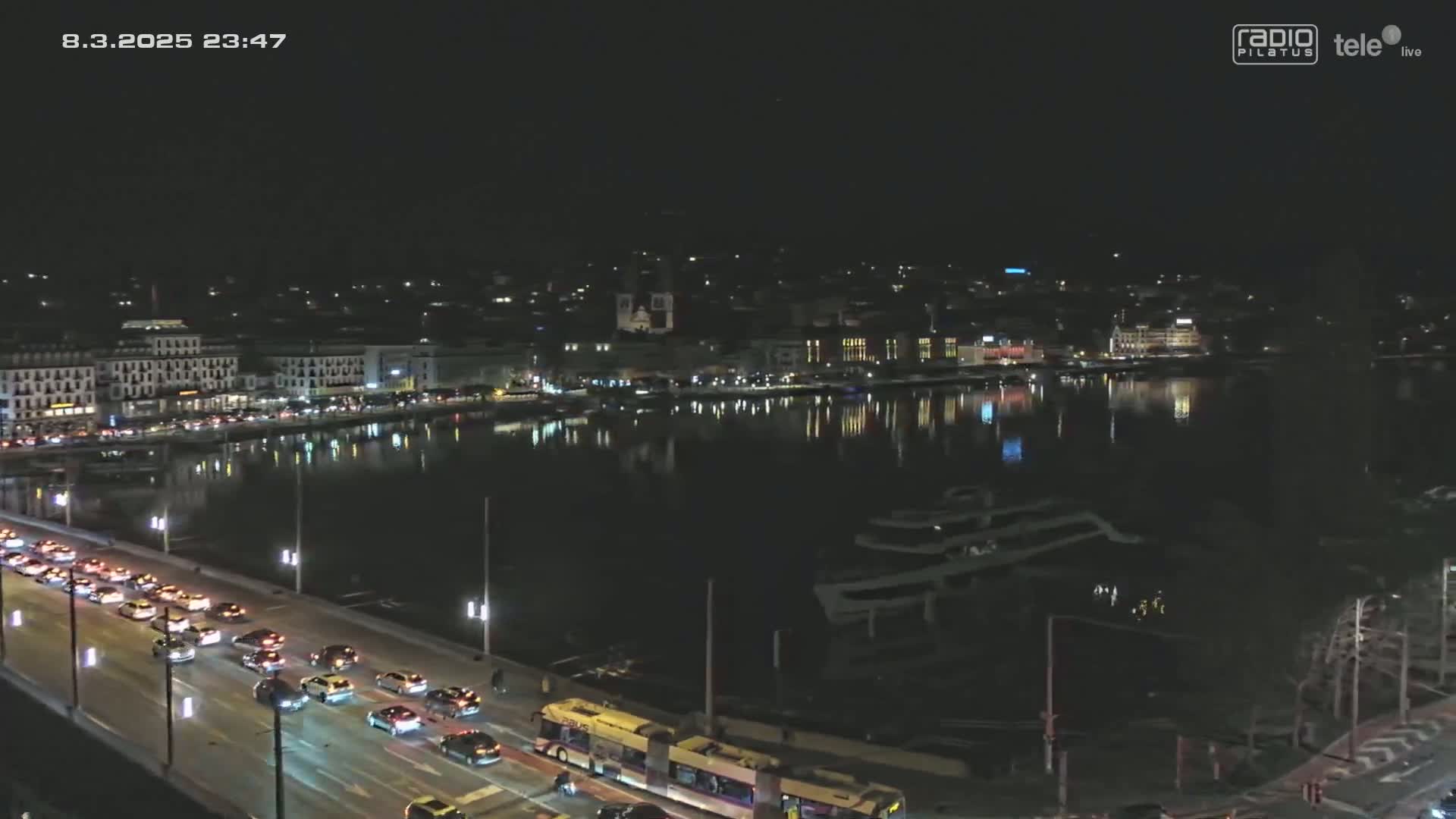 A nighttime, high-angle view shows a city's waterfront at night, with a bridge carrying heavy traffic leading to a calm body of water reflecting the city lights.
