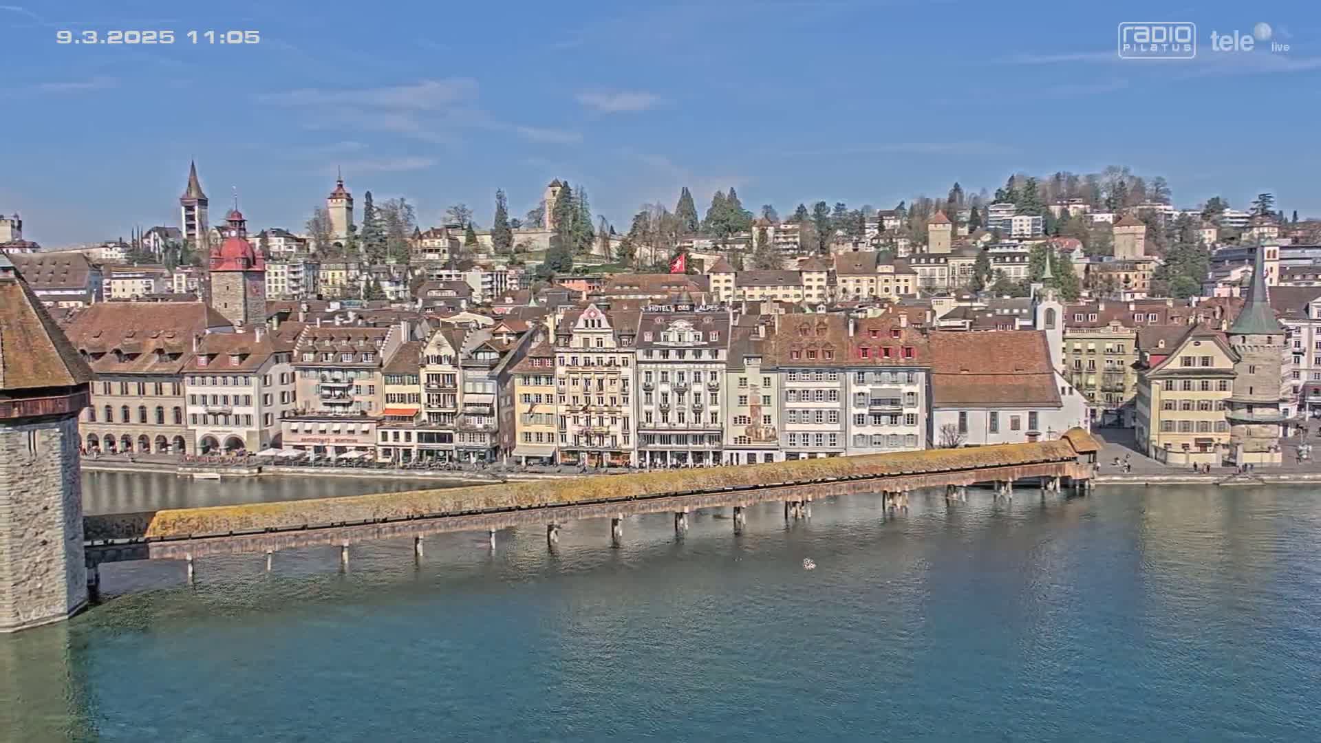 A picturesque waterfront town with many buildings, a covered bridge, and a calm body of water under a clear, sunny sky.