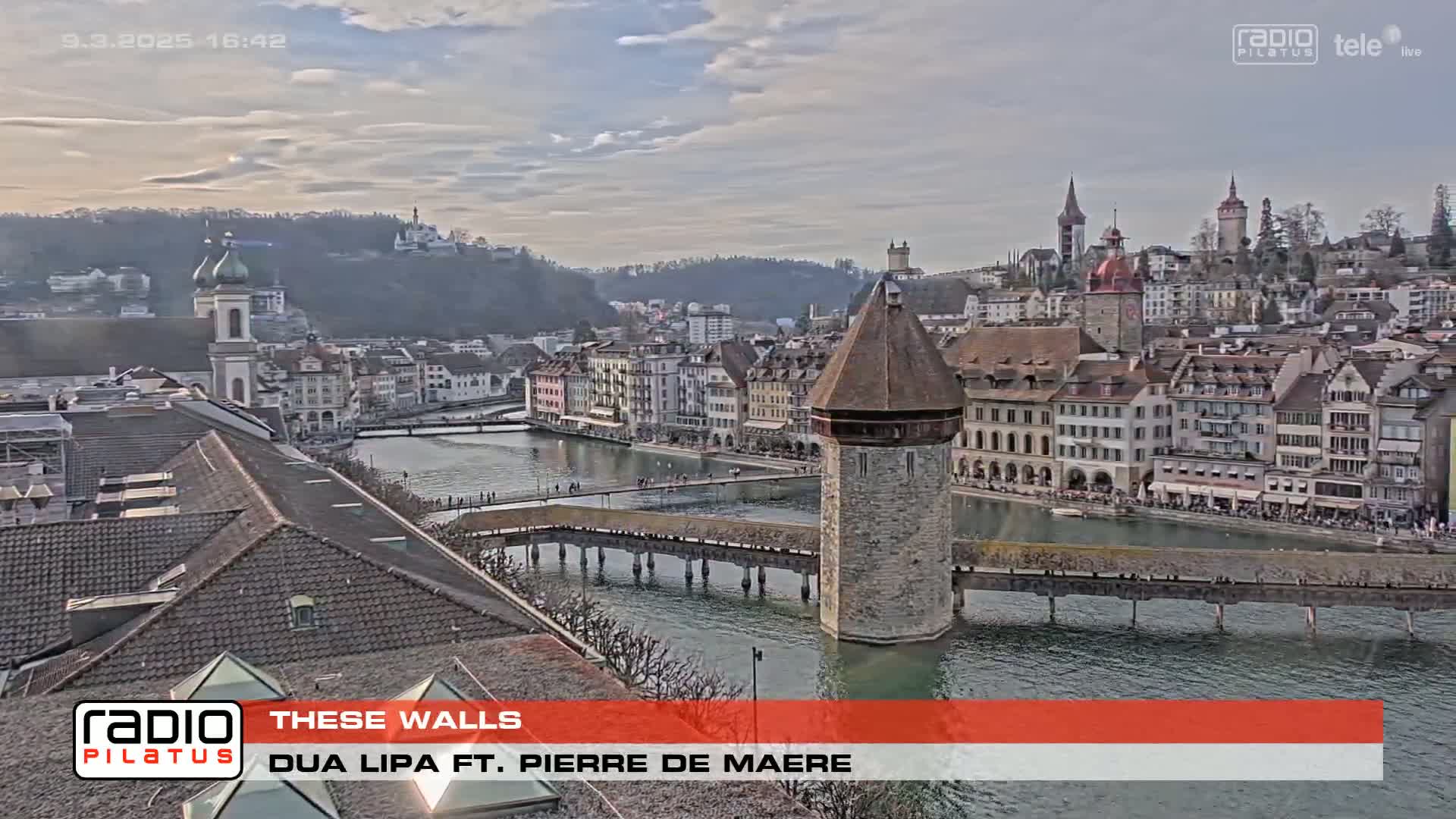 A quaint European town with a river running through it, under a partly cloudy sky, is viewed from a rooftop.