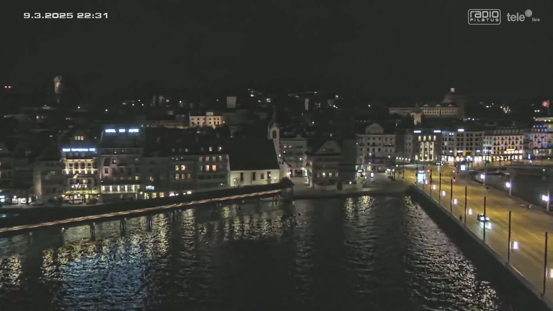 A nighttime, aerial view of a city waterfront shows a dark body of water reflecting the lights of buildings along the shore, with a road and several vehicles visible on a quay.