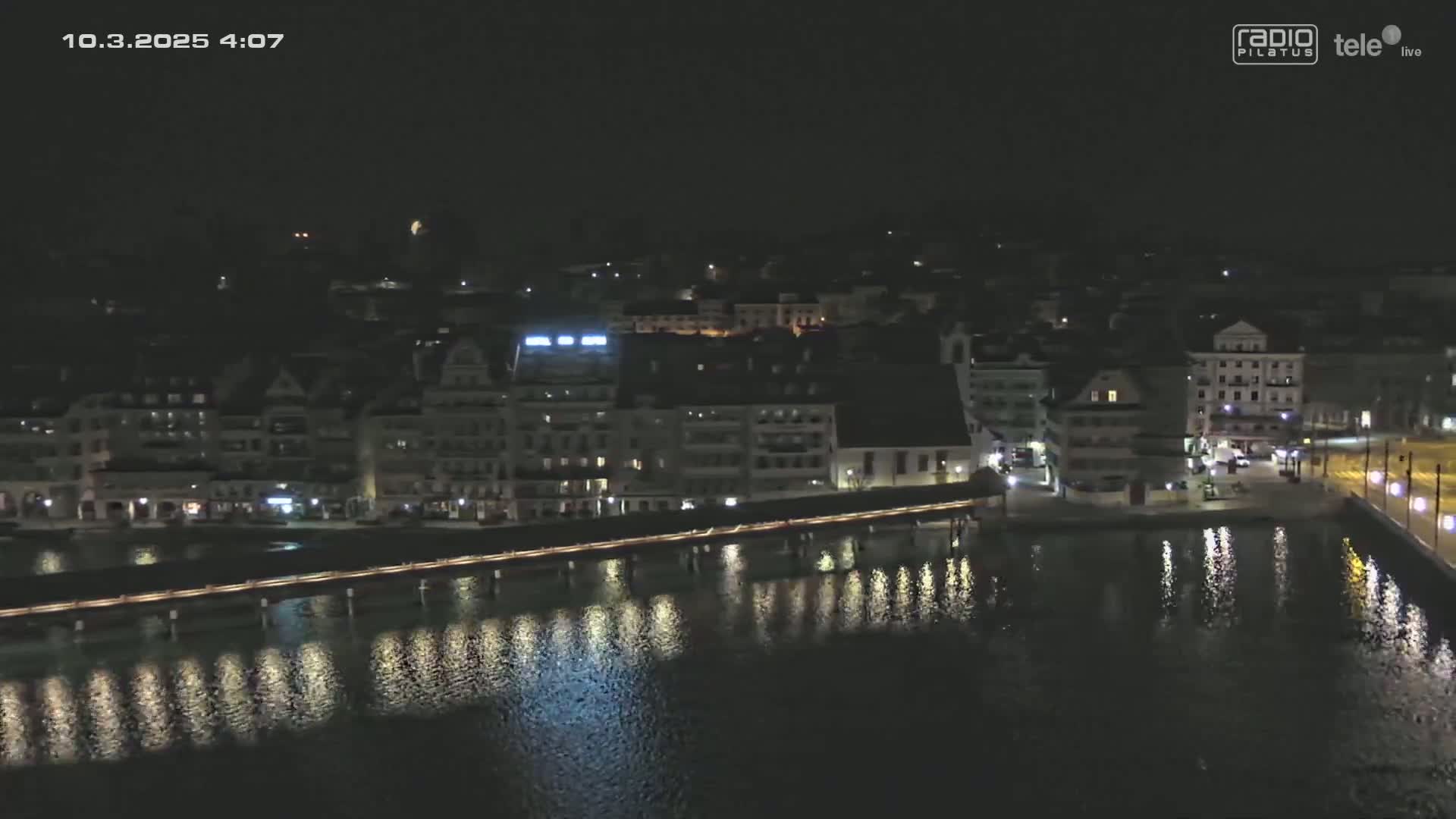 A nighttime aerial view of a city waterfront shows a bridge spanning a body of water reflecting the lights of buildings lining the shore.