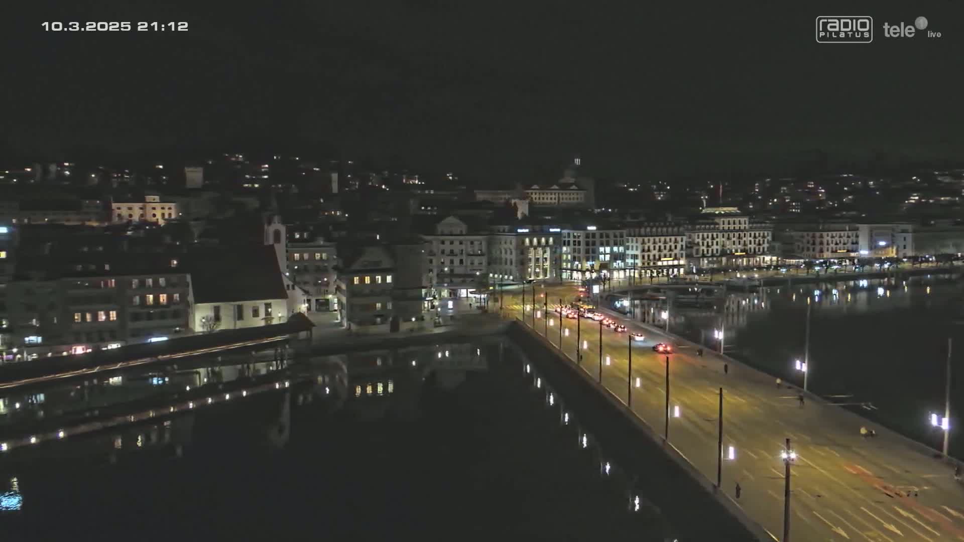 A nighttime, aerial view of a city beside a body of water shows a bridge with cars driving on it, buildings lining the waterfront, and their lights reflecting on the dark water.