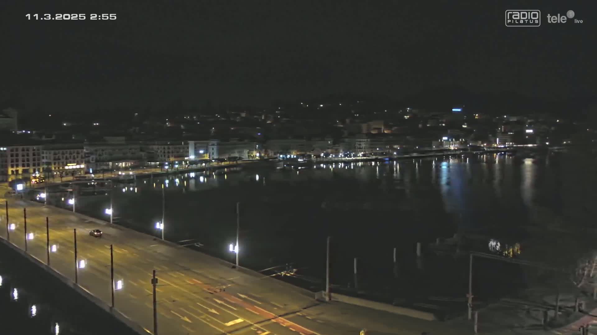A nighttime, high-angle view shows a city's waterfront at night, with a bridge and a calm body of water reflecting the city lights.