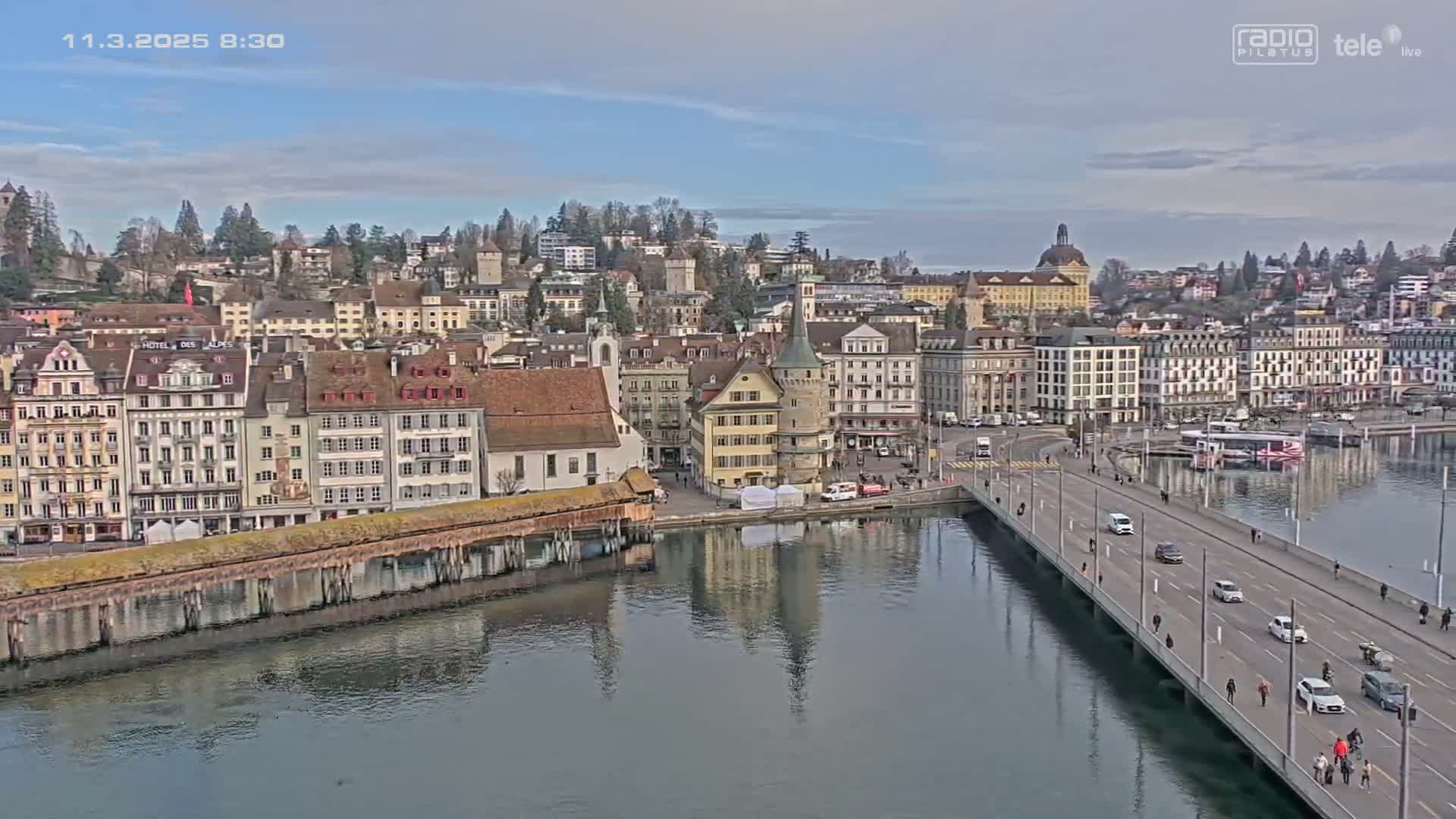 A bridge crosses a calm river, leading to a town of closely packed buildings under a mostly clear sky.