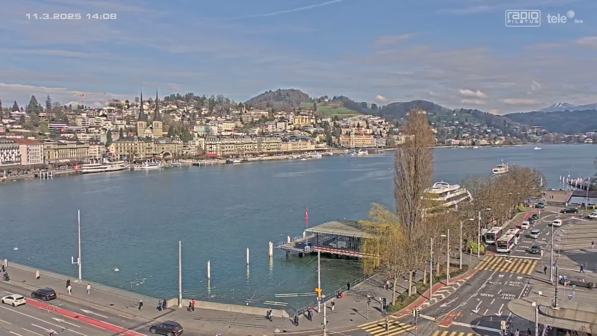 A waterfront city with buildings lining a calm lake under a mostly sunny sky is seen from an elevated viewpoint, with a road and pedestrian areas in the foreground.
