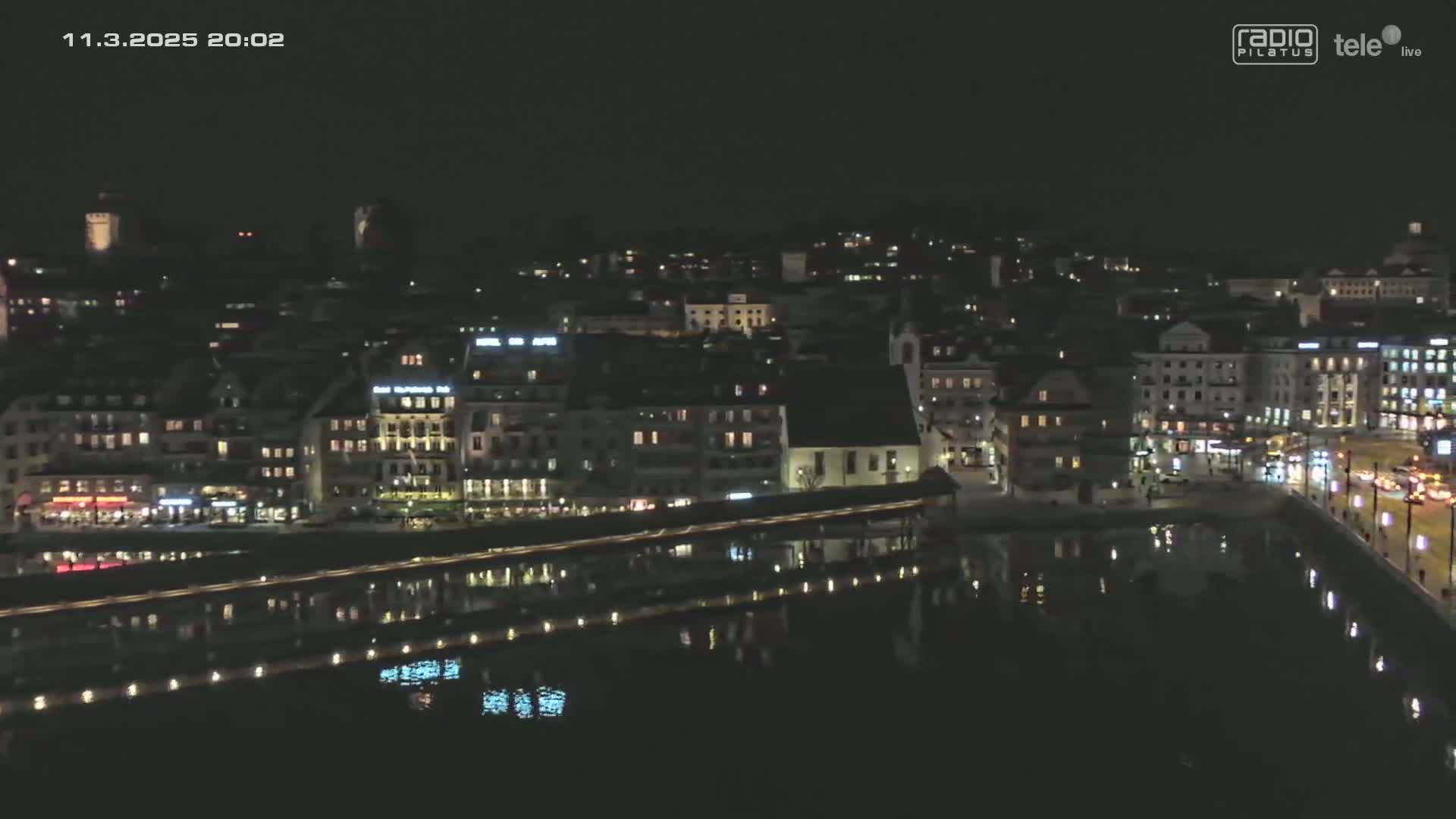 A nighttime view of a city with buildings illuminated along a body of water, reflecting the lights.