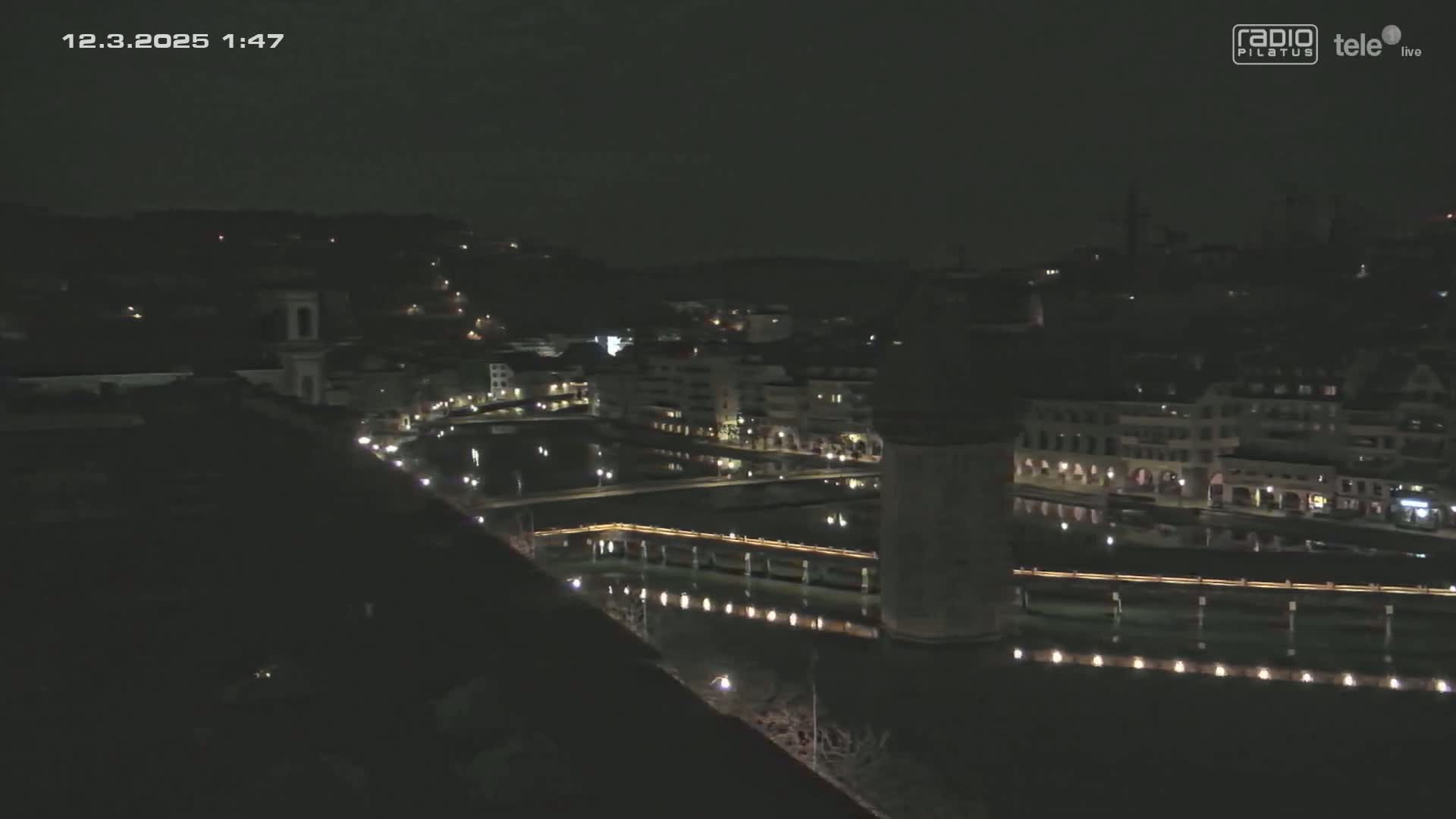 A nighttime, aerial view of a city with a river and bridge, showing buildings lit up along the water's edge under a dark sky.