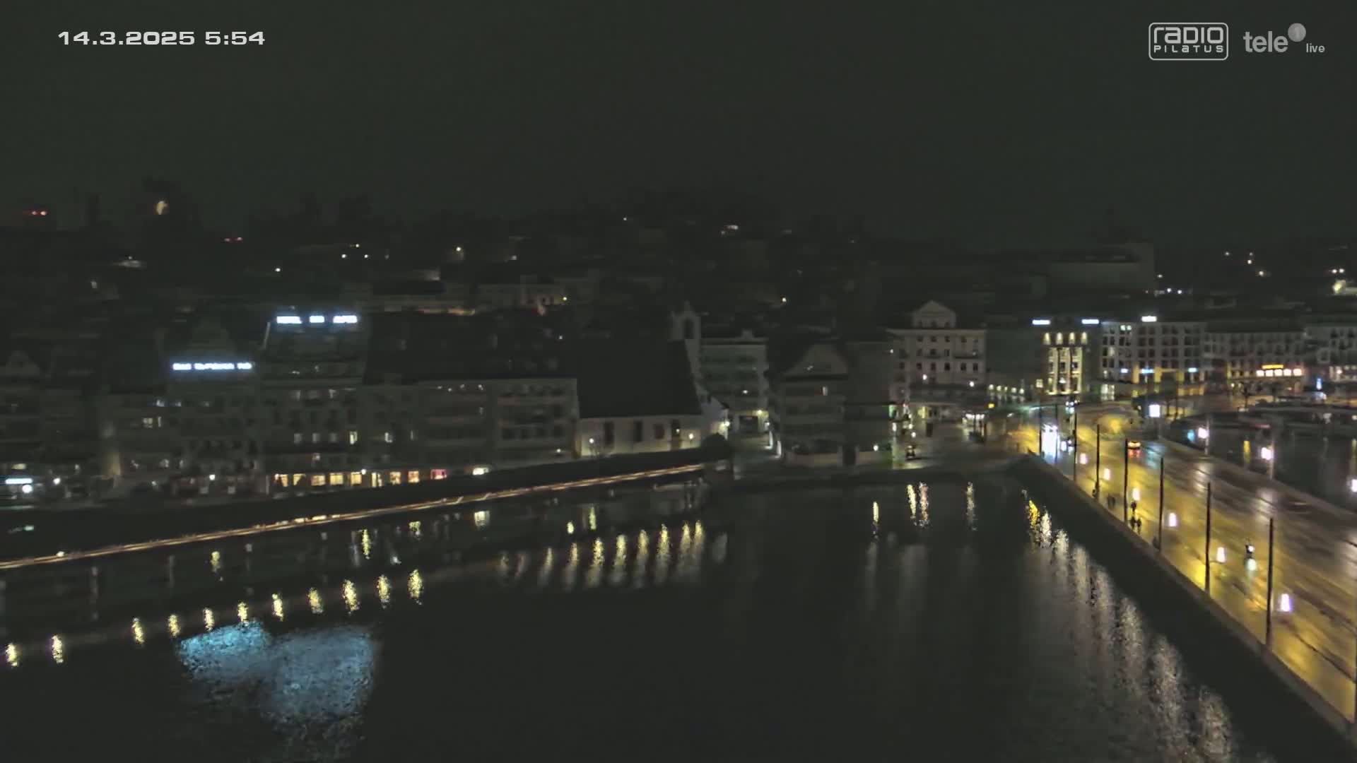 A nighttime aerial view shows a city's waterfront at night, with buildings lining a body of dark water reflecting city lights.