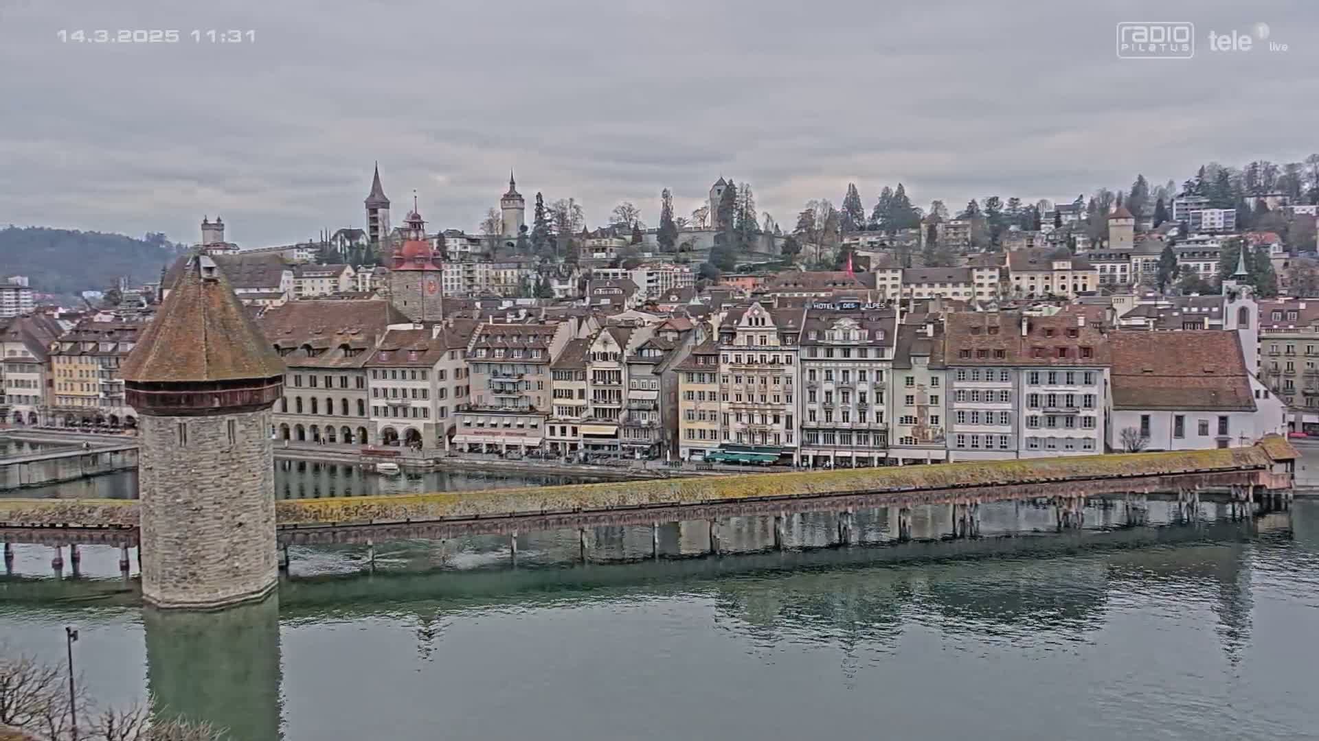 A stone water tower sits on a bridge spanning a river, with a cityscape of numerous buildings behind it under an overcast sky.