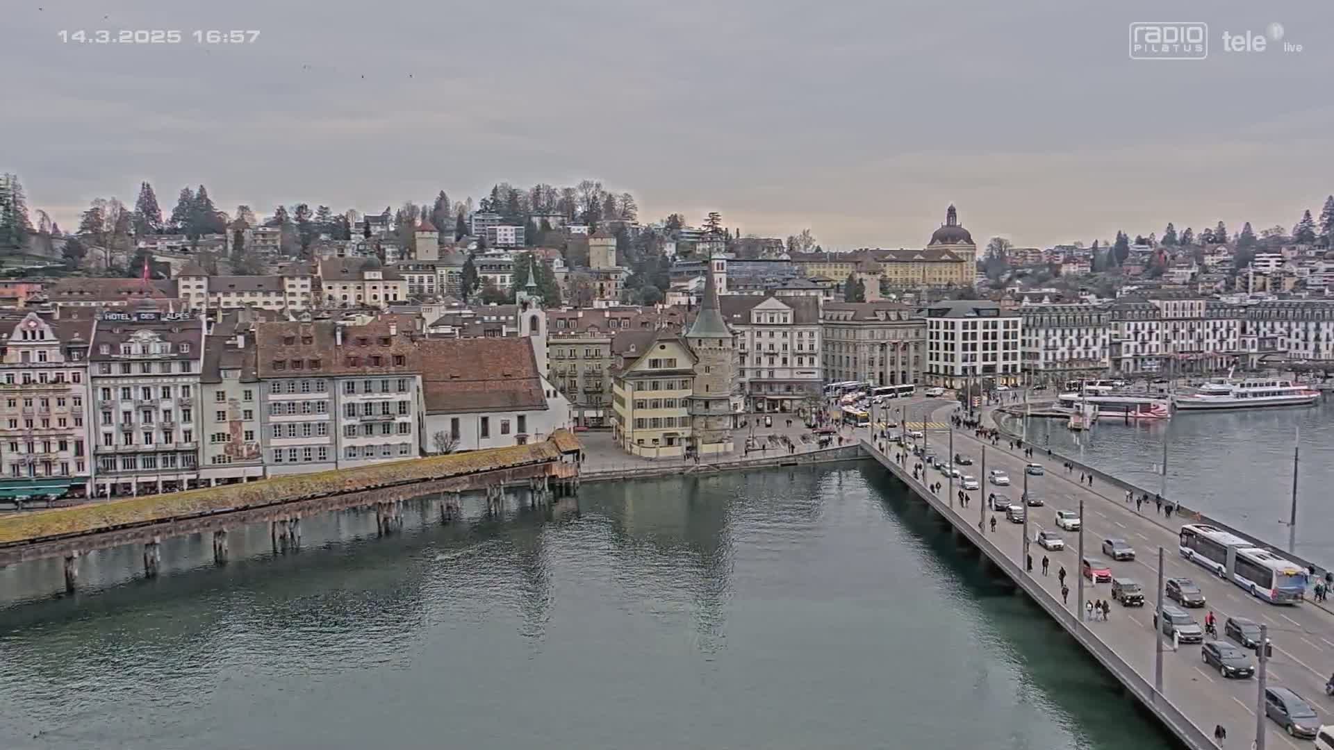 A bridge over a body of water with cars driving across it, and a town with many buildings visible in the background under an overcast sky.