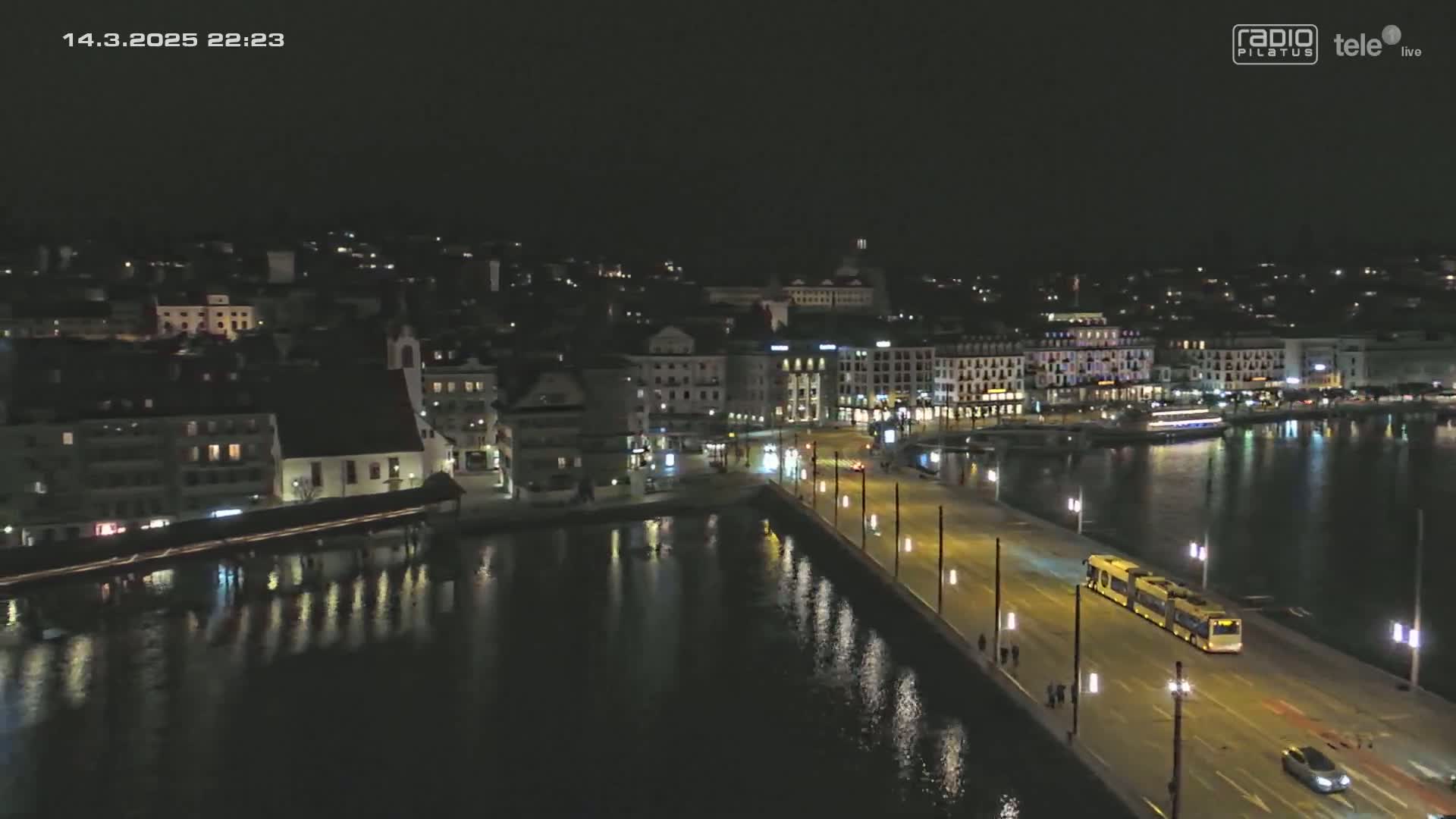 A nighttime aerial view of a city by the water, showing a bridge with a tram and illuminated buildings along the waterfront under a clear, dark sky.