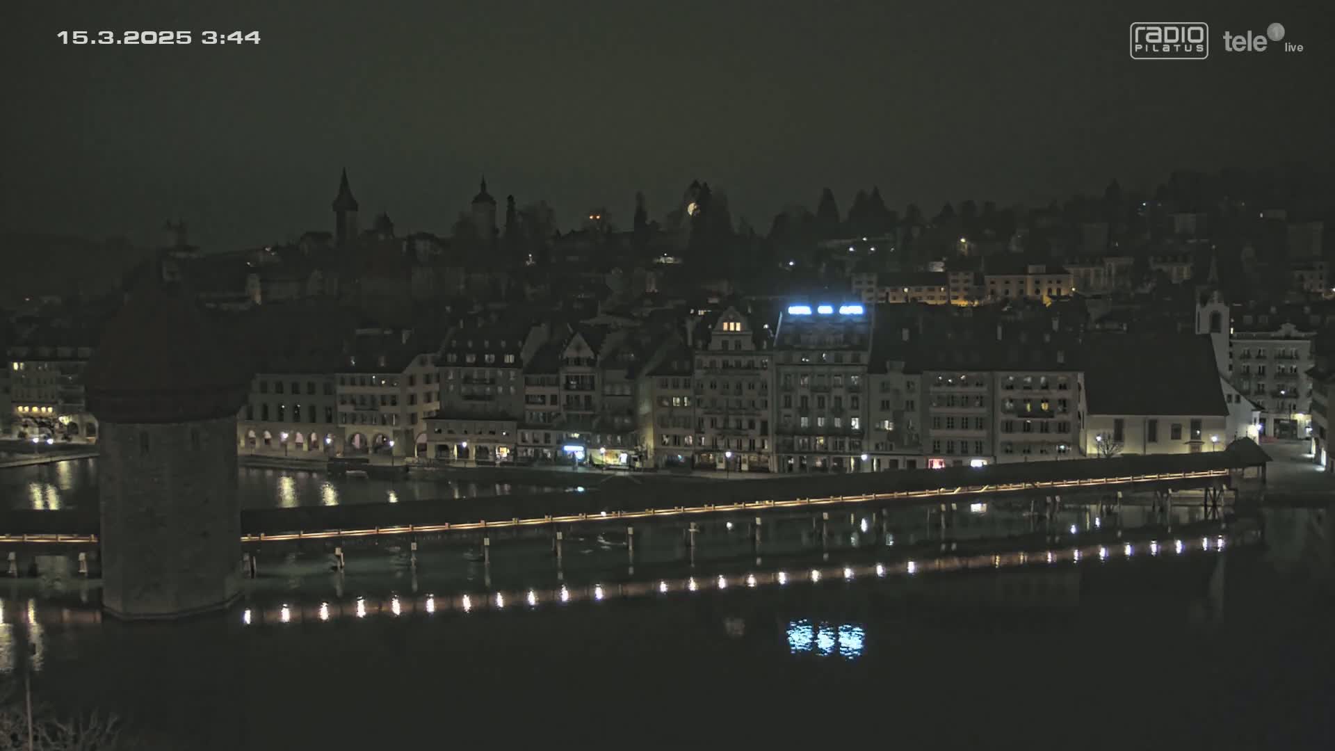 A nighttime view of a city with a bridge spanning a dark body of water, illuminated by lights.