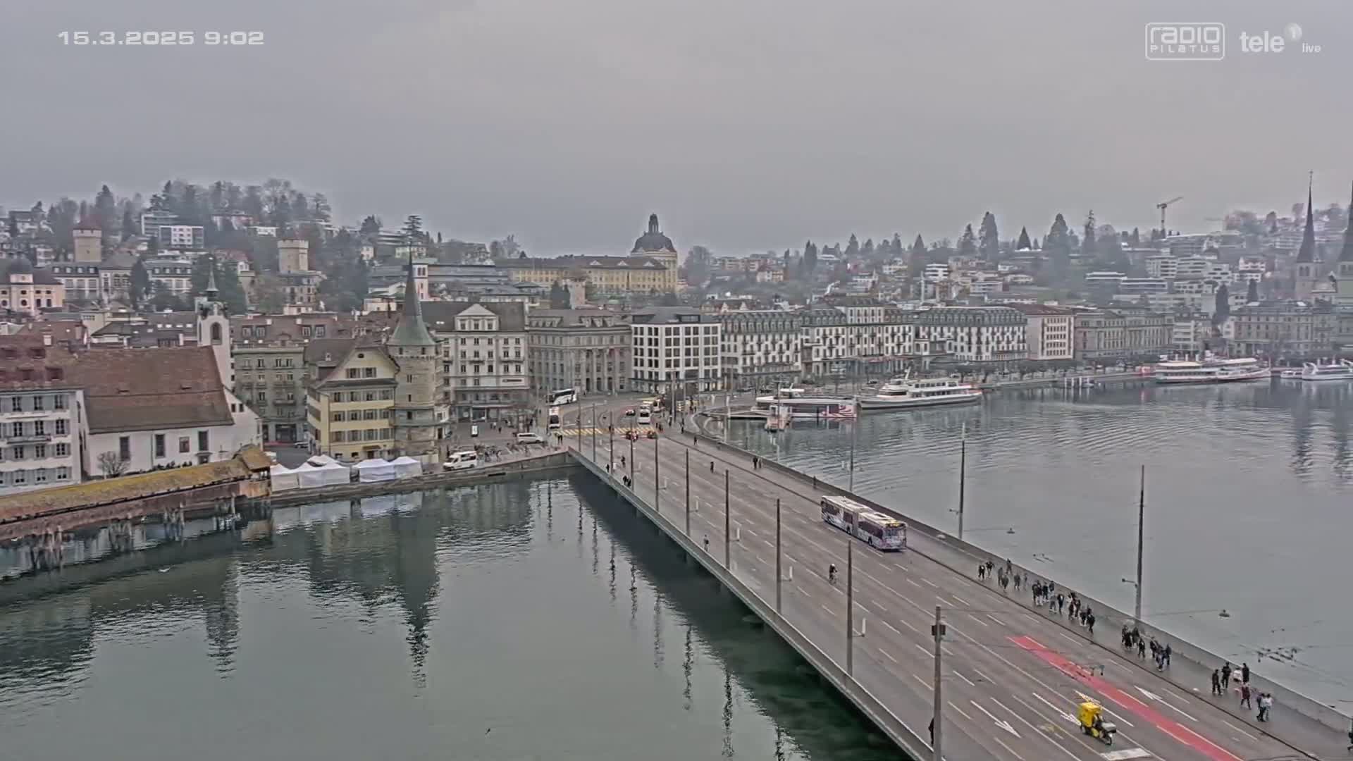 A long bridge over a calm river leads to a city of European-style buildings under an overcast sky.