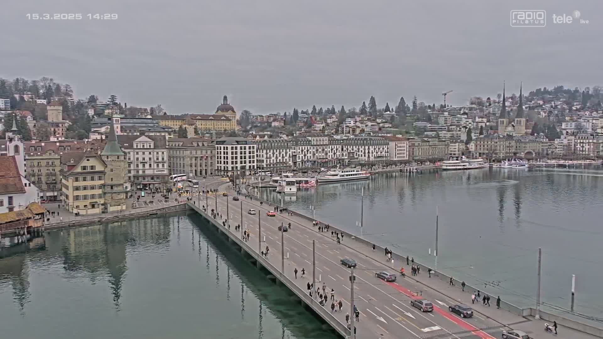 A long bridge stretches across calm, dark green water, lined with pedestrians and cars, leading to a city of old and new buildings under a gray, overcast sky.