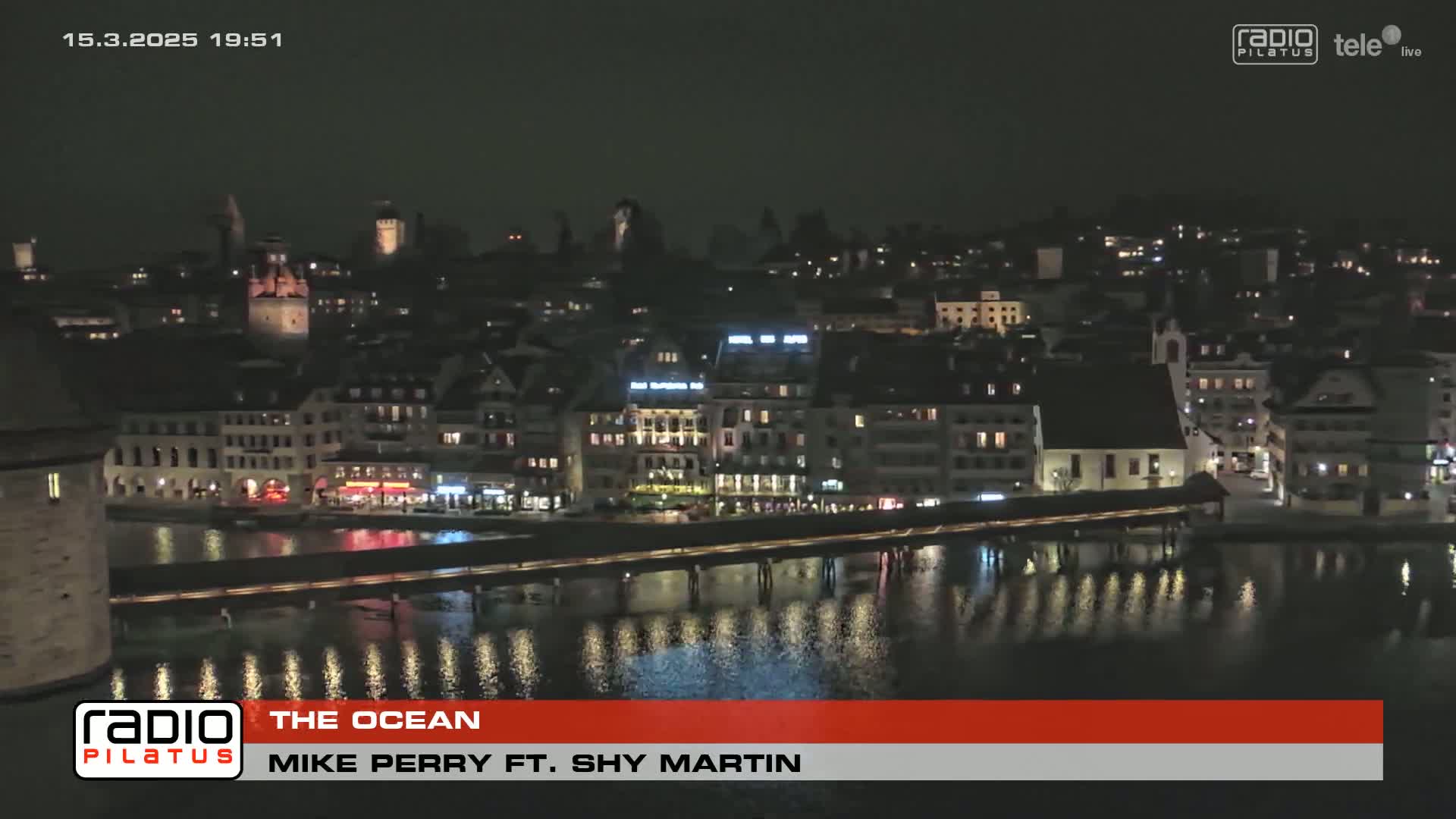 A nighttime view of a city illuminated along a waterfront, with a bridge and calm water reflecting the lights.