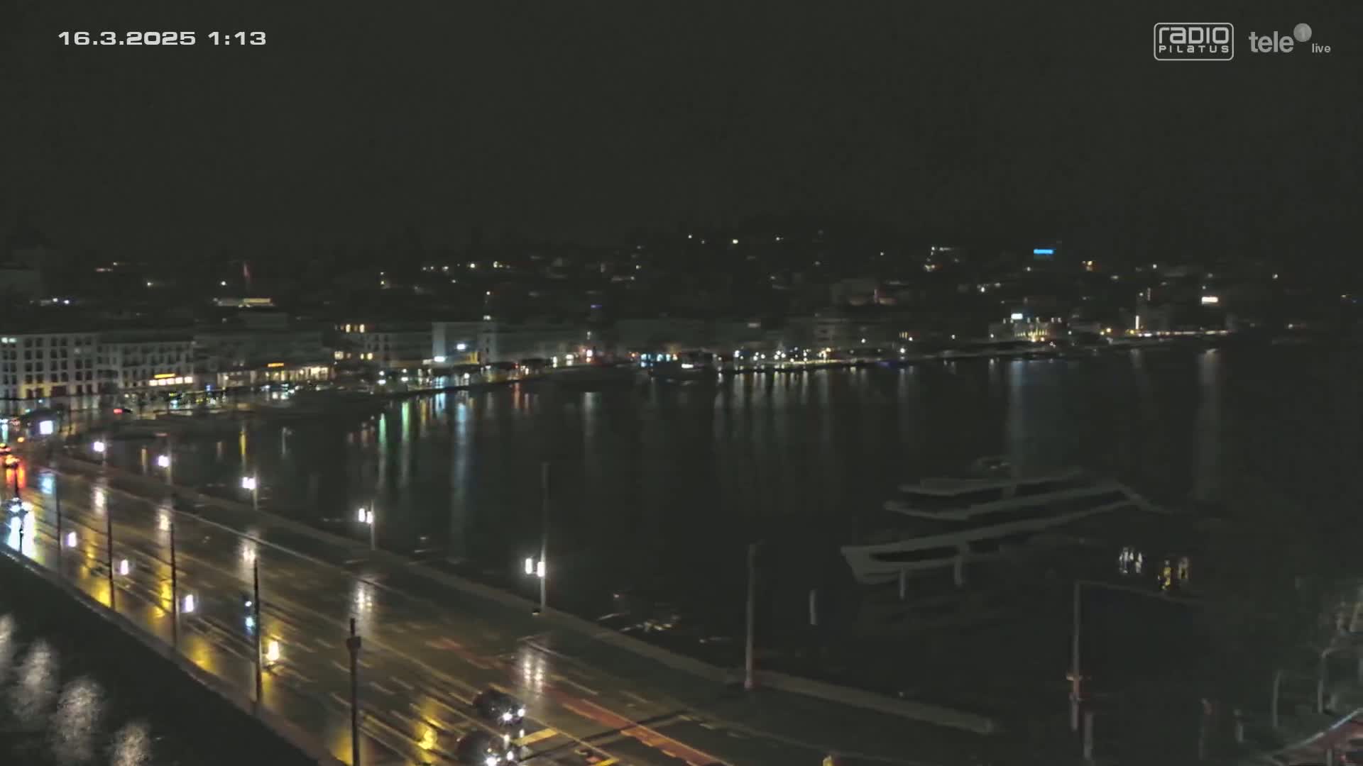 A nighttime view of a city's waterfront, showing a wet roadway with cars driving on a bridge overlooking a calm body of water with docked boats, under dark, possibly rainy conditions.