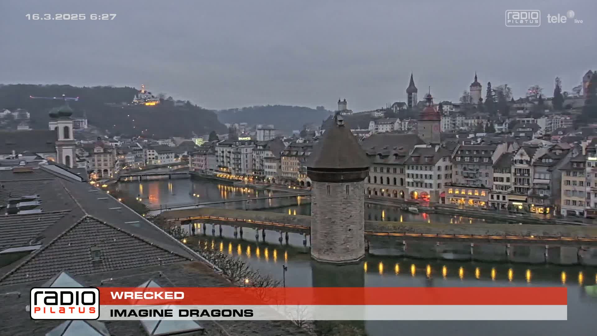A twilight view of a European city, featuring a river with lights reflecting on its surface, historic buildings lining the waterfront, and a stone tower in the middle of a bridge.