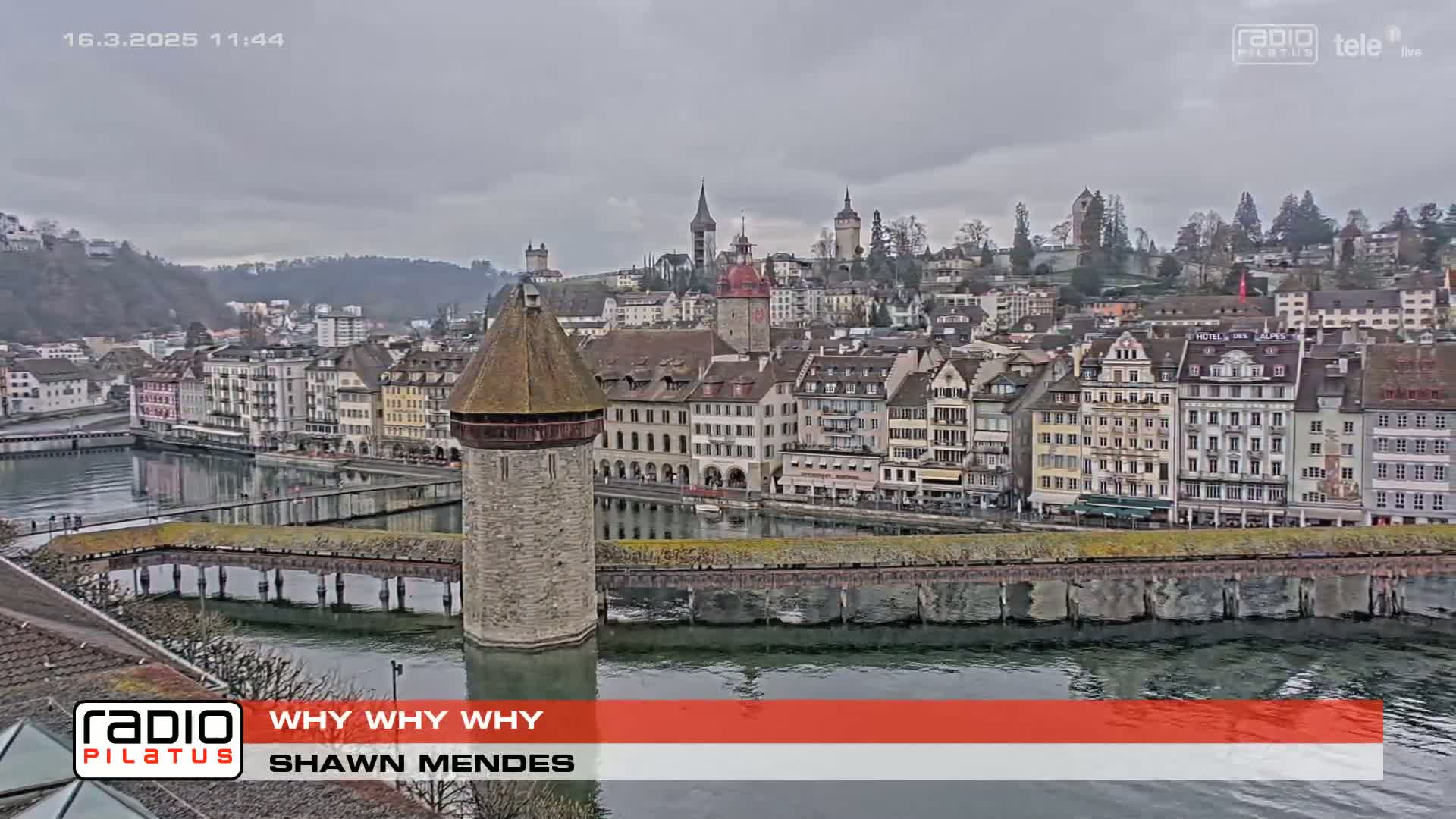 A gray, overcast day reveals a European town situated along a river, featuring a stone tower on a bridge and numerous buildings lining the waterfront.