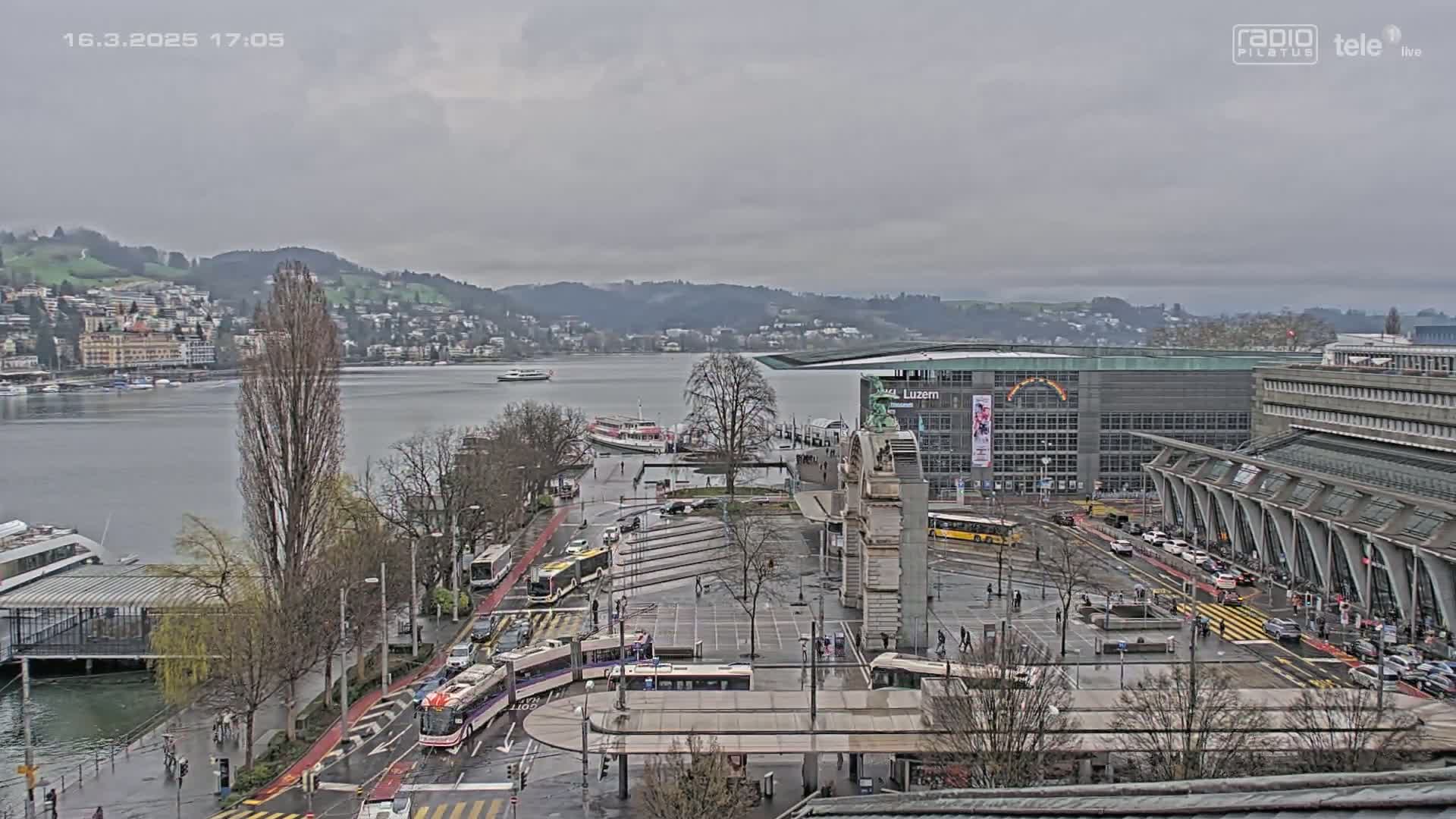 A wet, overcast city scene shows a harbor with boats, a large square with buses and trams, and buildings along a waterfront, backed by hills.