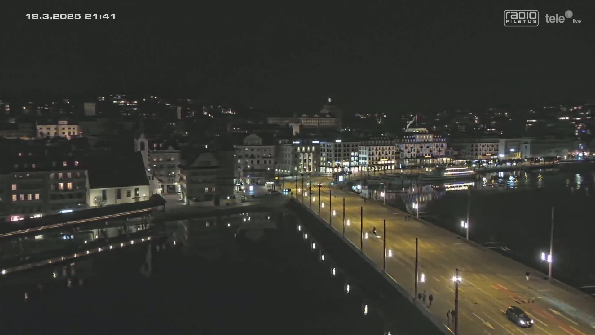A nighttime aerial view of a city beside a body of water shows a bridge illuminated with streetlights and sparsely-populated by vehicles and pedestrians.