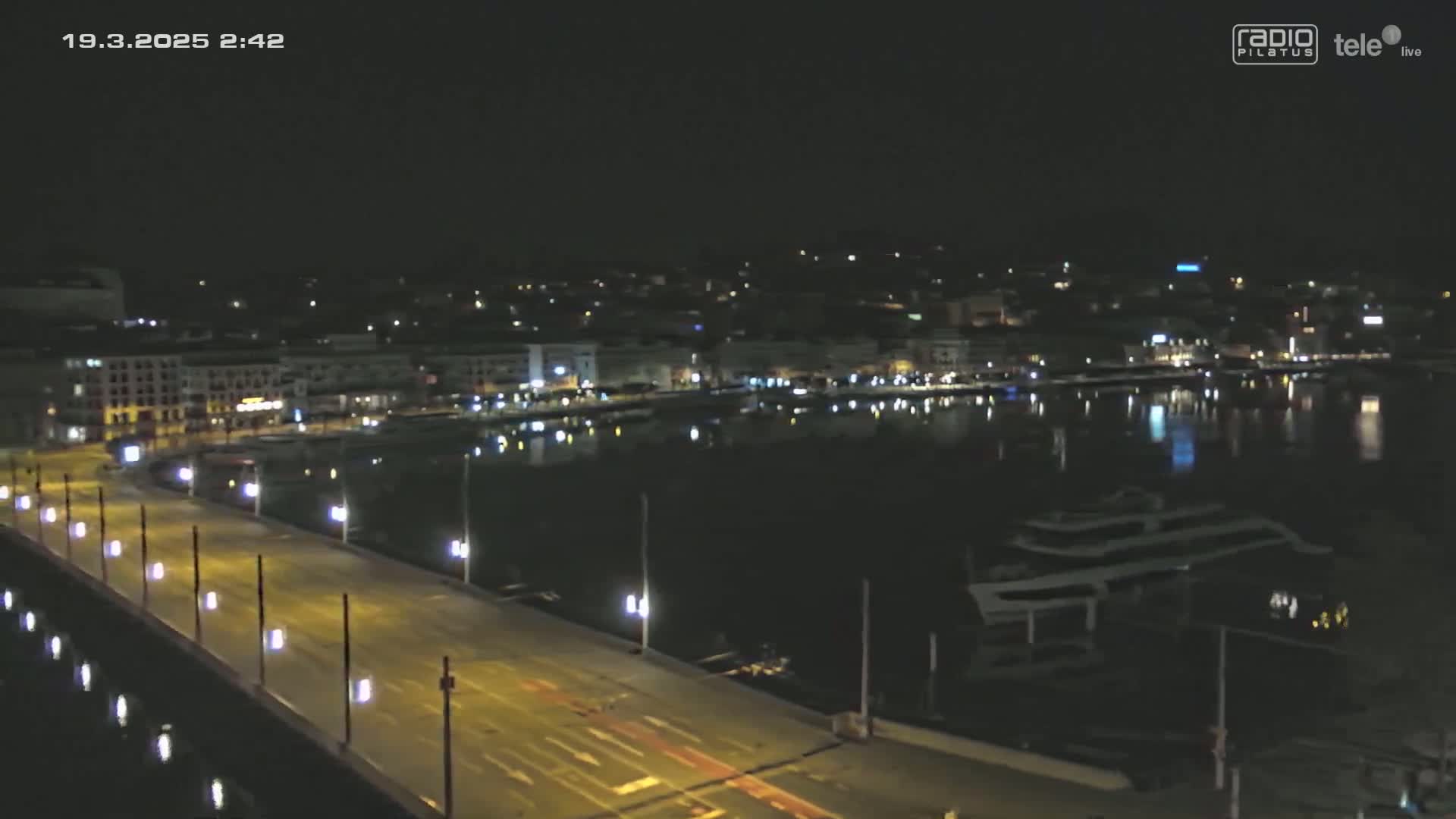 A nighttime, high-angle view shows a brightly lit bridge over dark water, with a city's lights reflecting in the water and a large vessel moored nearby.