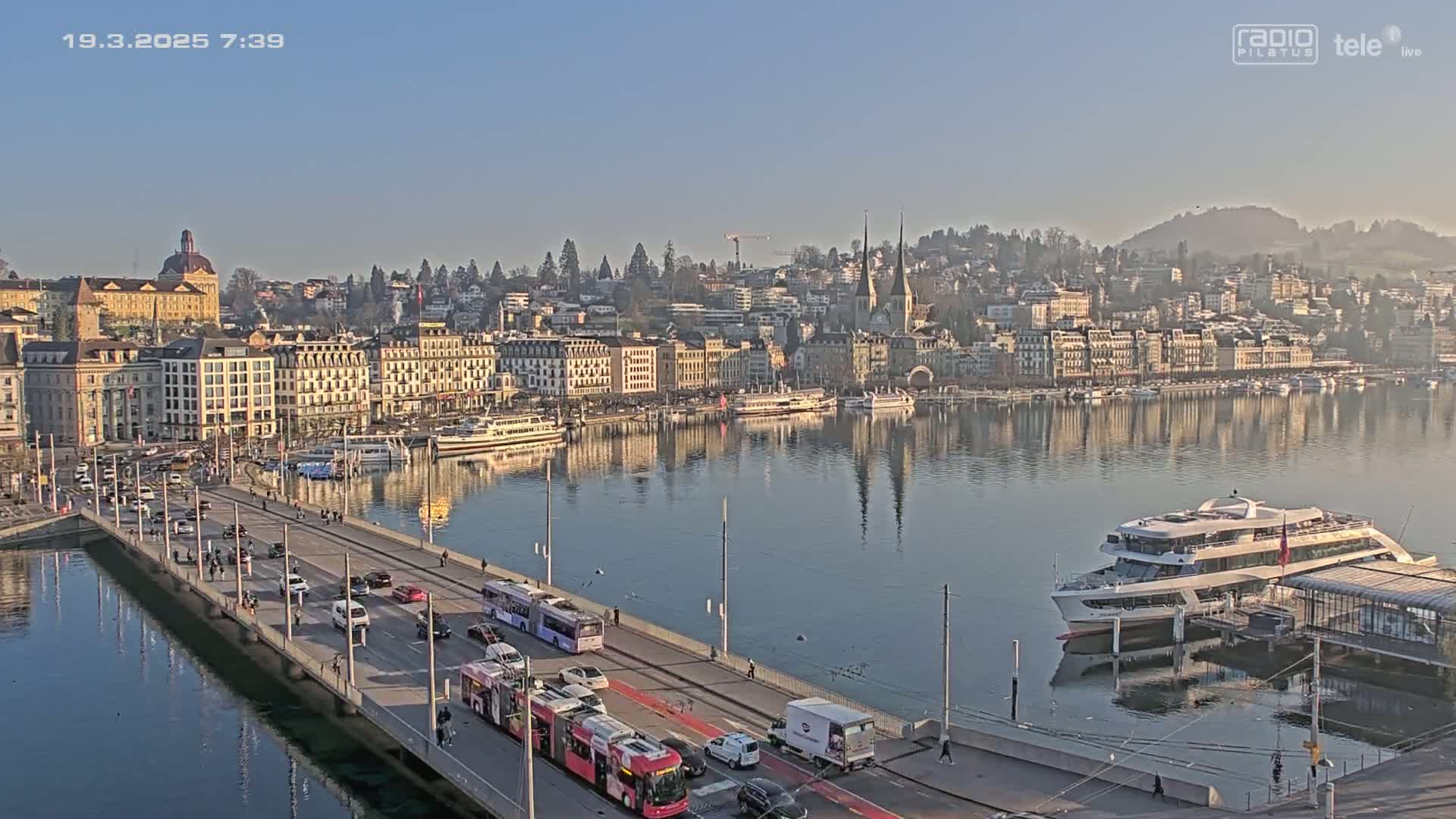 A bridge with traffic crosses a calm lake reflecting a city of buildings under a clear, sunny sky.