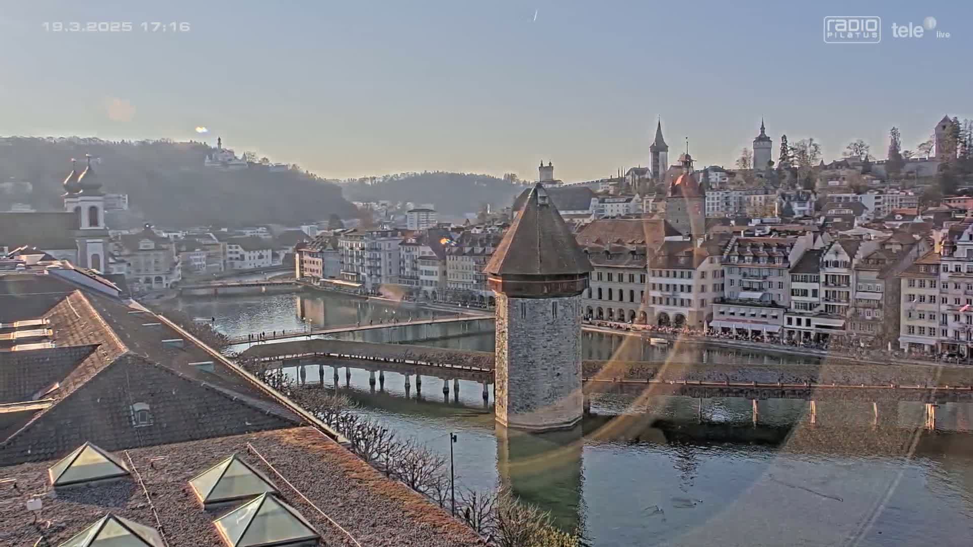 A waterside European city with a stone tower at the center of a bridge under a clear, sunny sky.