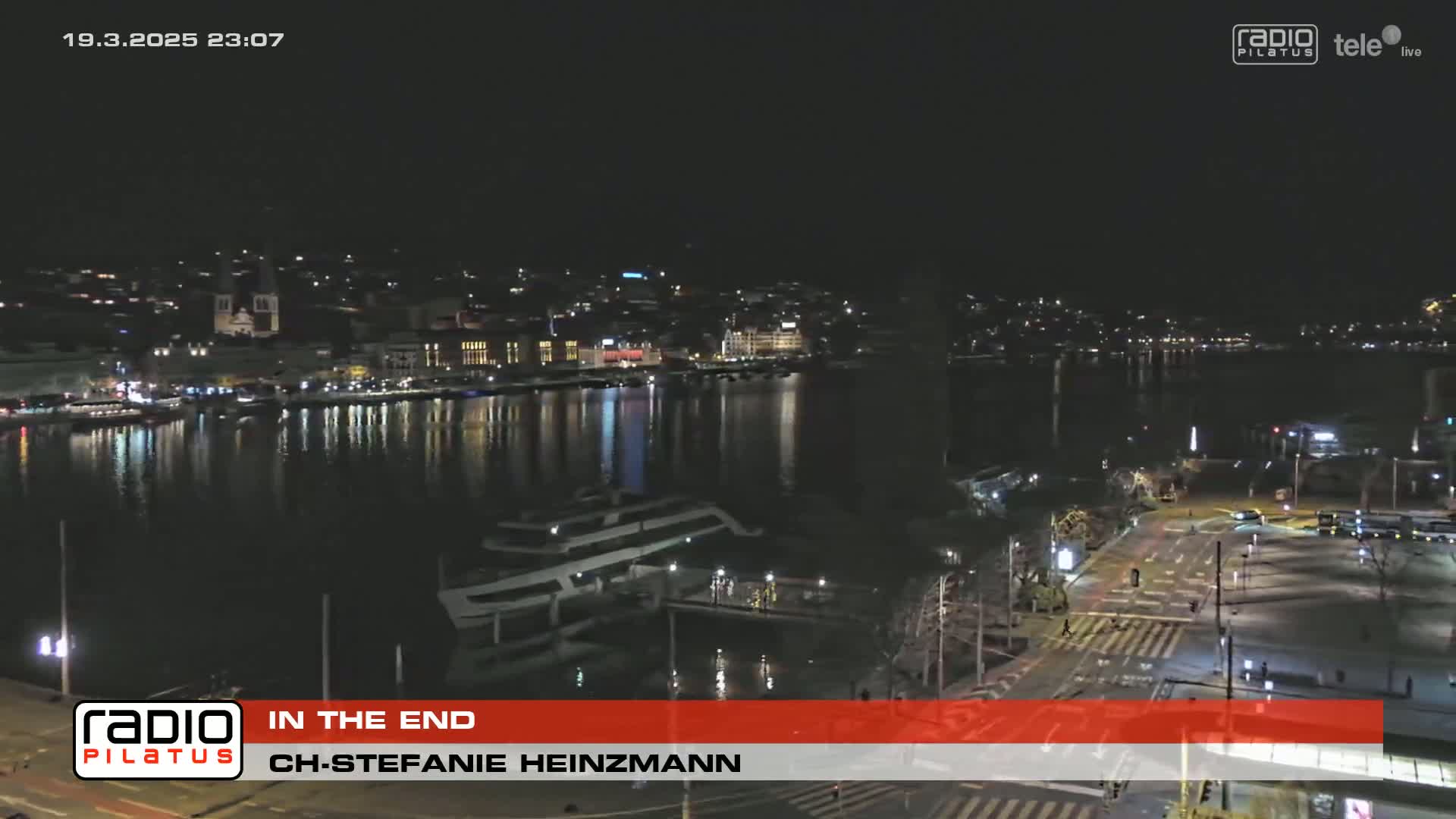 A nighttime view of a city waterfront with calm water reflecting city lights, and a roadway with sparsely parked cars.
