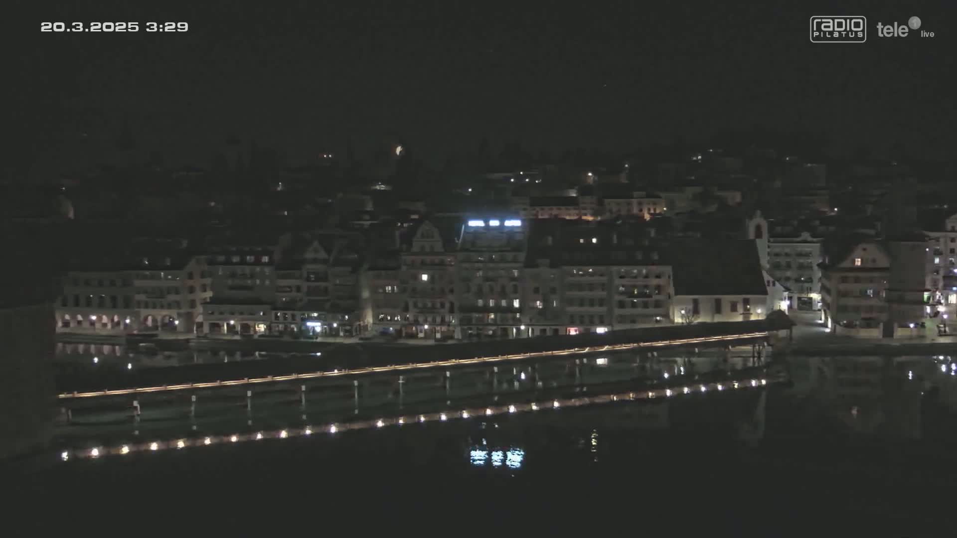 A nighttime, high-angle view shows a town's waterfront at night, with a long bridge reflecting in dark water.