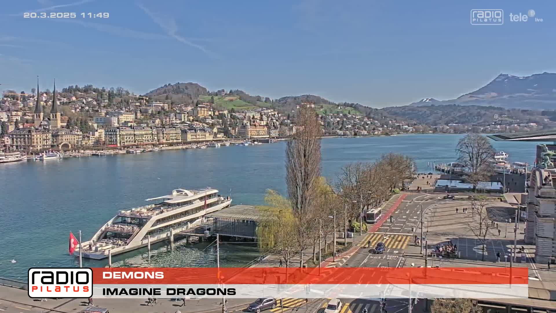 A sunny day overlooking a waterfront city with a large boat docked near a paved area, and mountains in the background.
