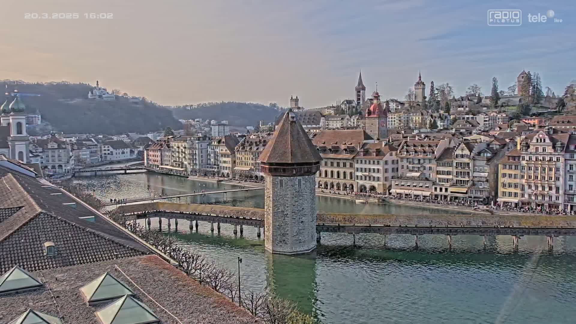 A stone tower sits in the middle of a covered bridge spanning a river, with a collection of buildings lining the riverbanks under a partly cloudy sky.