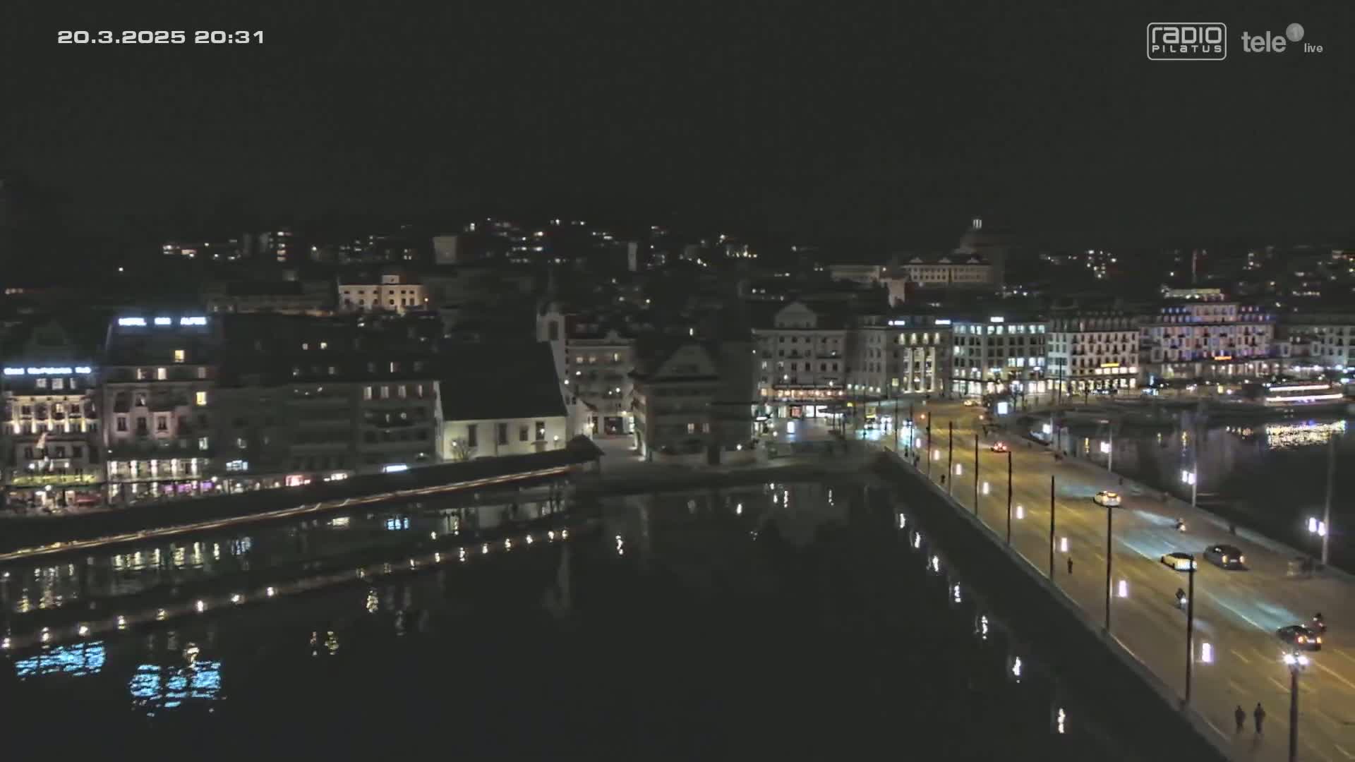 A nighttime aerial view shows a city's waterfront with buildings lining a calm body of water and a bridge with cars driving on it.